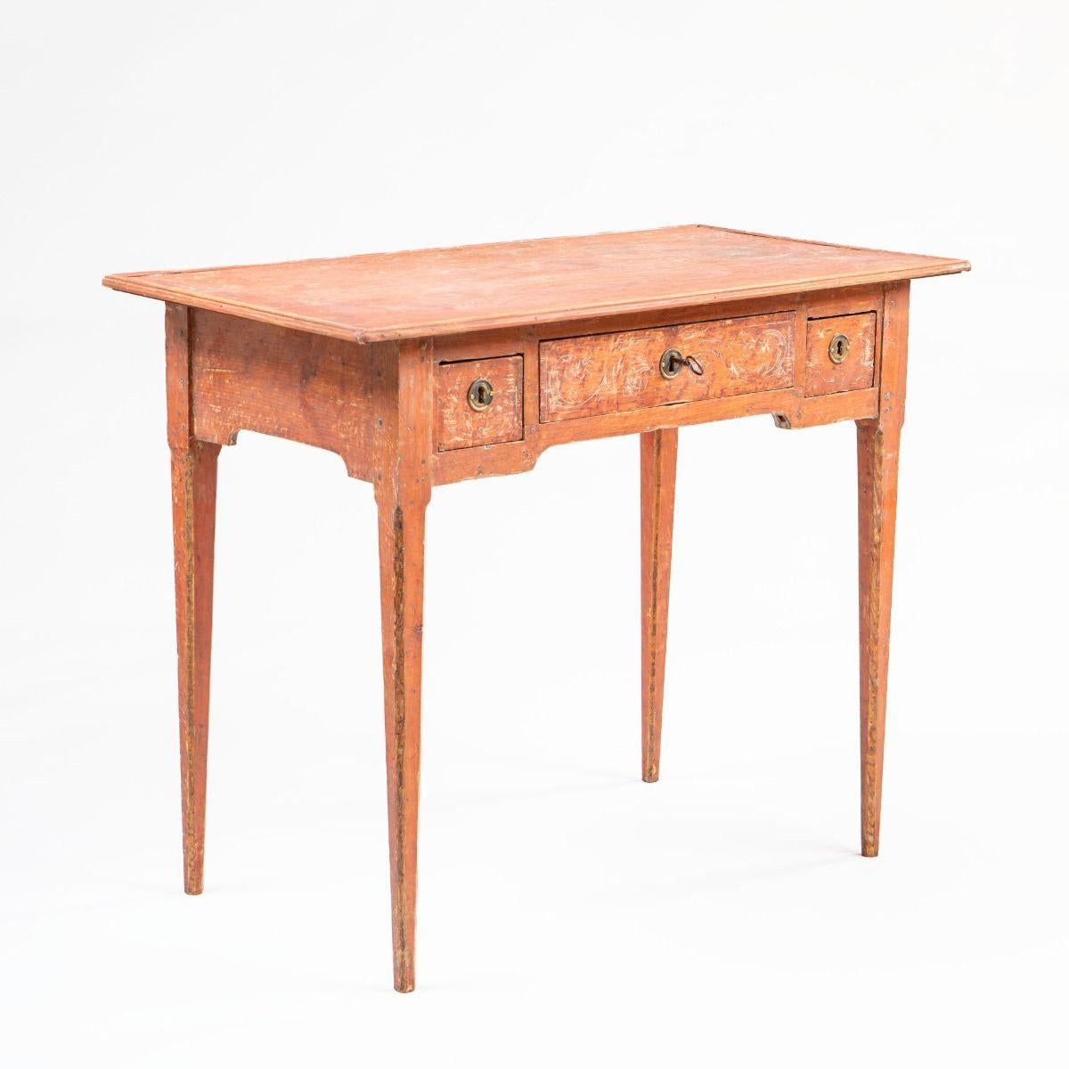 Belle et raffinée table Gustavienne / Louis XVI en pin, soigneusement grattée pour révéler sa peinture d'origine de couleur corail avec une belle patine.

Le plateau présente un bord profilé et un tablier avec trois tiroirs, dont un large tiroir