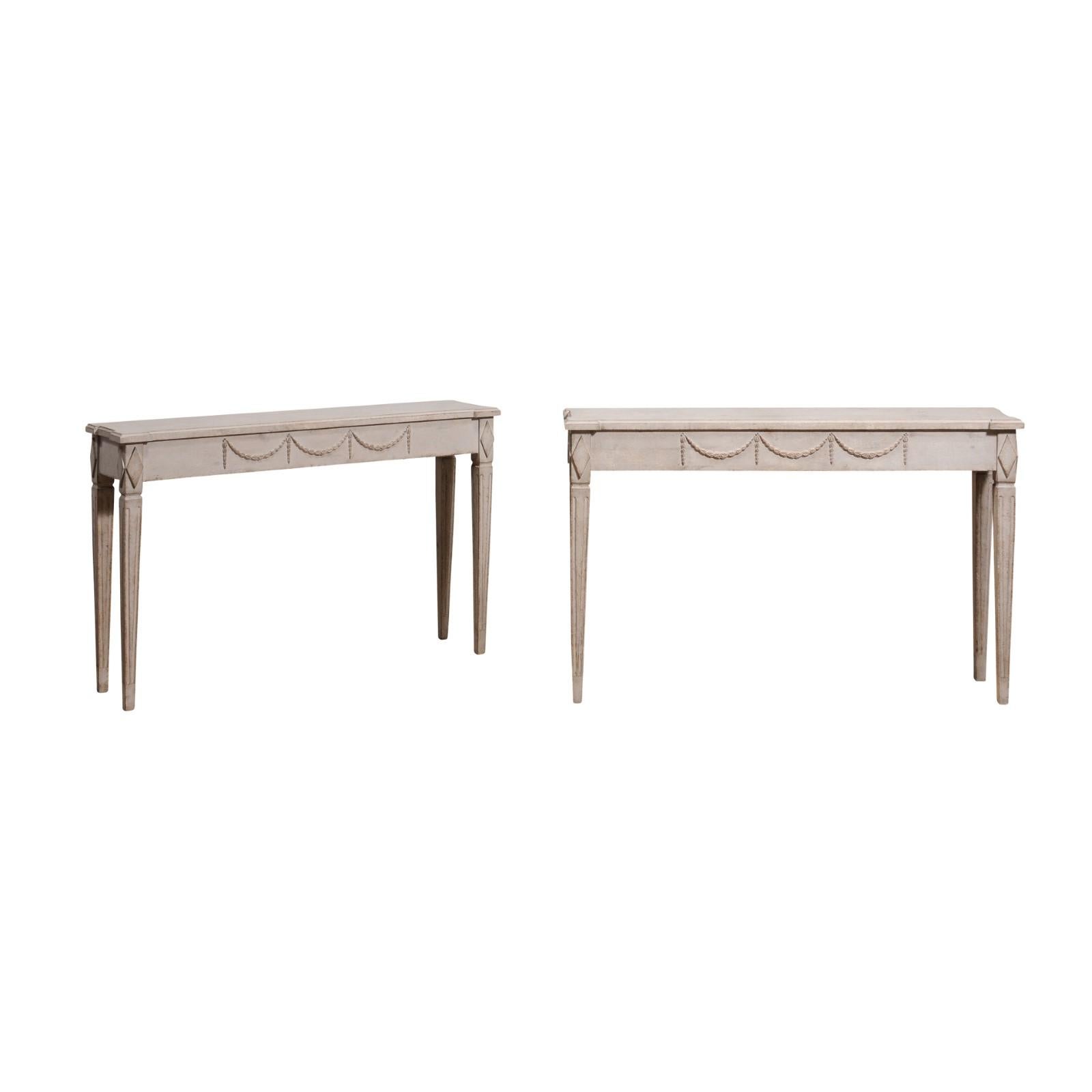 A pair of Swedish Gustavian period console tables from circa 1810 with light gray painted finish, carved garlands and diamond motifs, and tapered fluted legs. This exquisite pair of Swedish Gustavian period console tables, dating back to circa 1810,
