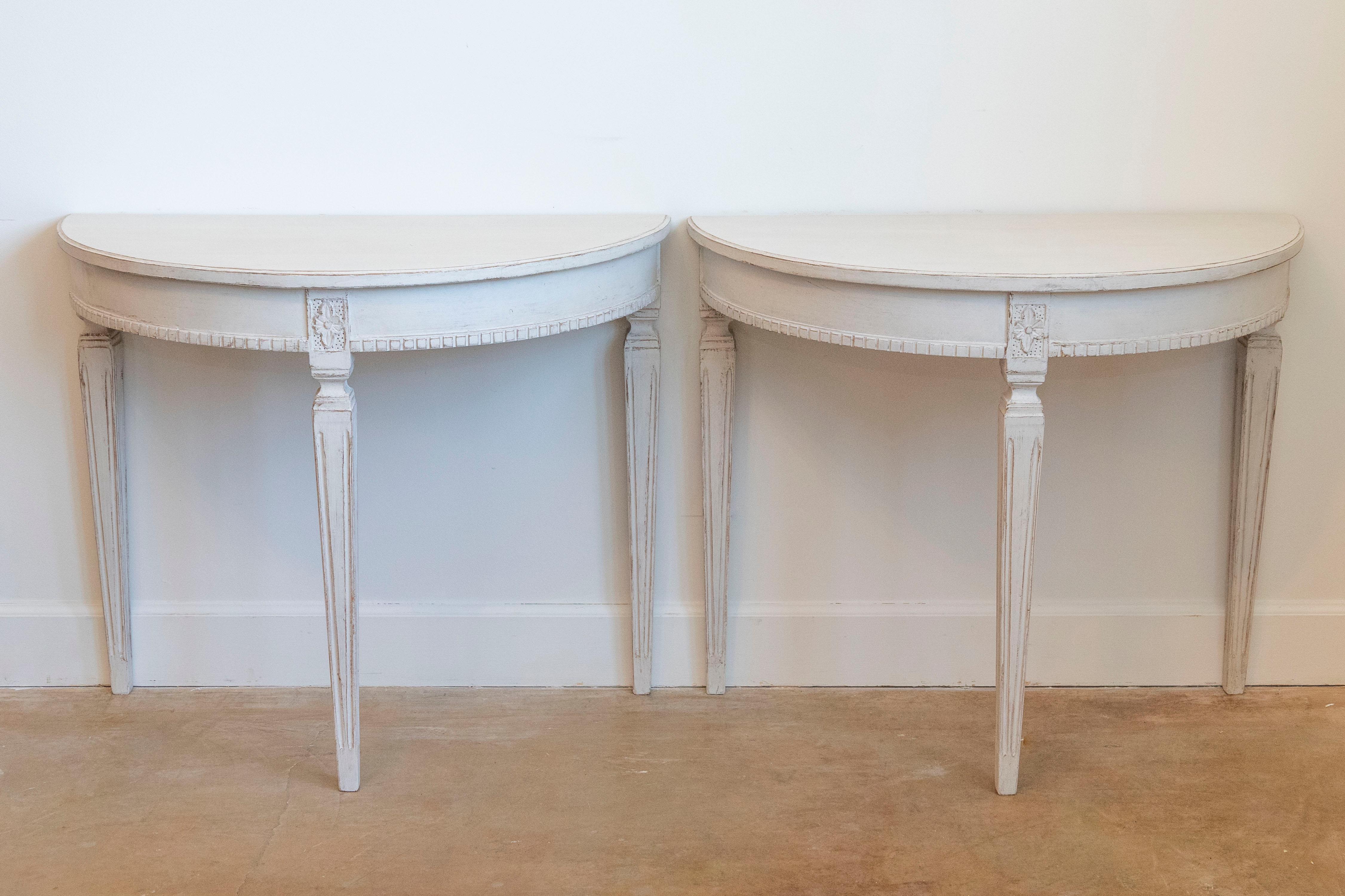 A pair of Swedish Gustavian style light gray / cream painted demi-lune console tables from circa 1880 with fluted tapered legs, dentil molding and carved rosettes on the knees. This pair of Swedish Gustavian style demi-lune console tables, crafted