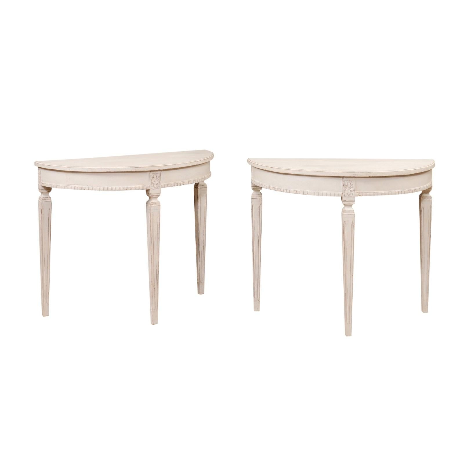 A pair of Swedish Gustavian style light gray / cream painted demi-lune console tables from circa 1880 with fluted tapered legs, dentil molding and carved rosettes on the knees. This pair of Swedish Gustavian style demi-lune console tables, crafted