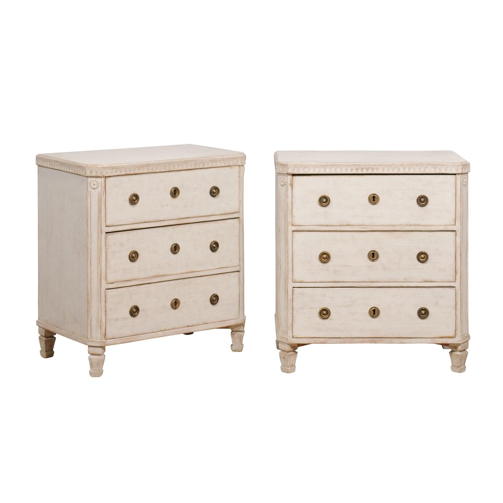 A pair of Swedish Gustavian style chests from circa 1880 with light gray / cream painted finish, three drawers each, carved friezes and tapered feet. A symphony of Swedish elegance, this pair of Gustavian style chests, dating from circa 1880, stands