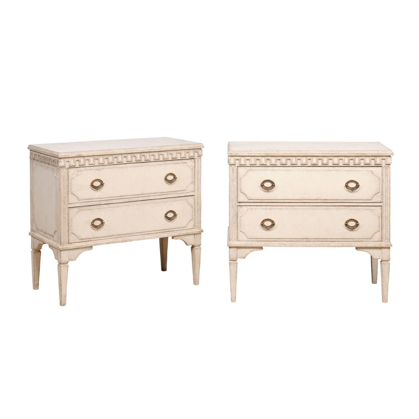A pair of Swedish Gustavian style chests from the 19th century with antique off white / cream painted finish, carved Greek Key frieze, two drawers and tapered legs. Step into the epitome of 19th-century Swedish design with this resplendent pair of