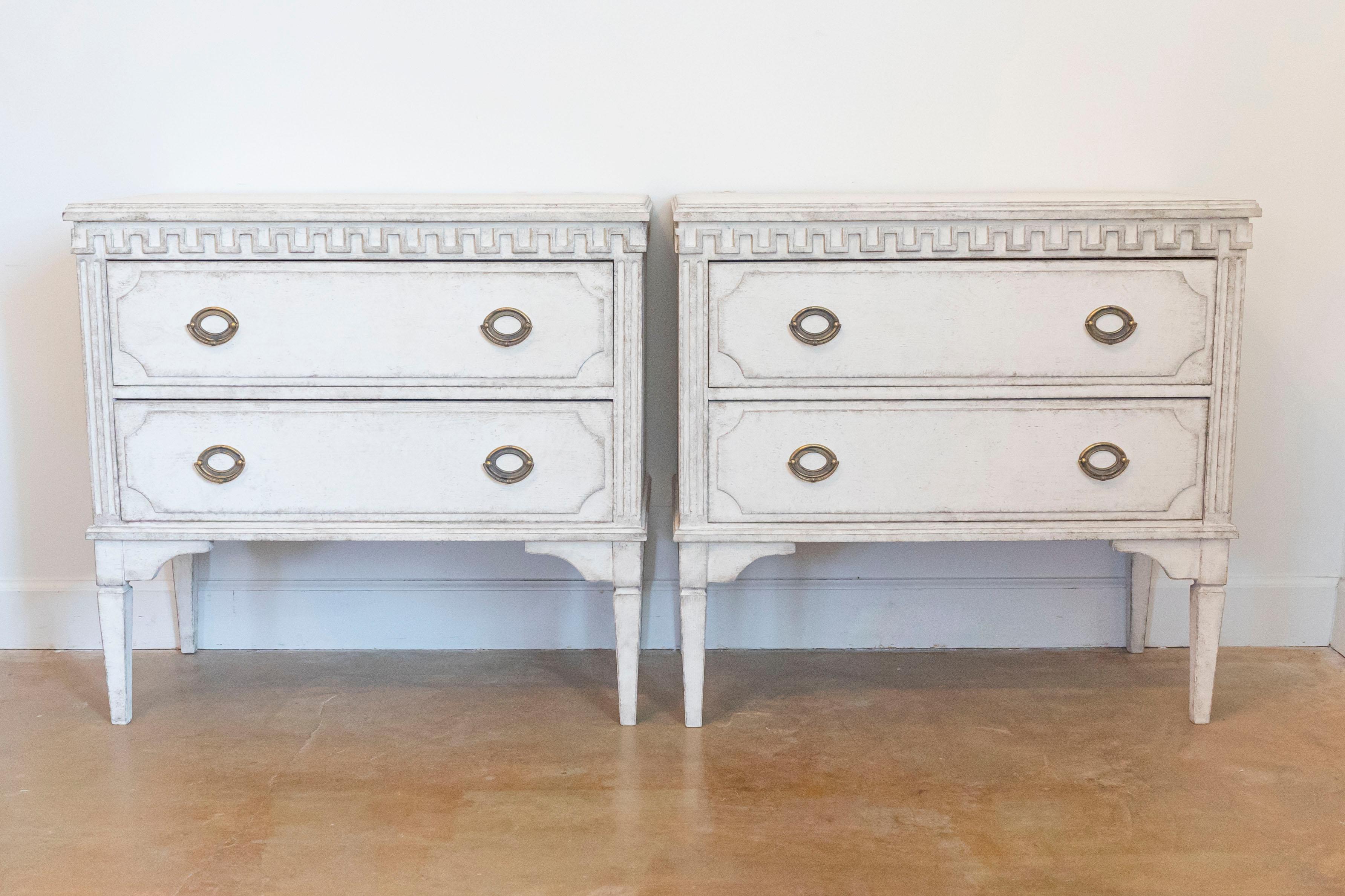 A pair of Swedish Gustavian style chests from the 19th century with antique off white / cream painted finish, carved Greek Key frieze, two drawers and tapered legs. Step into the epitome of 19th-century Swedish design with this resplendent pair of