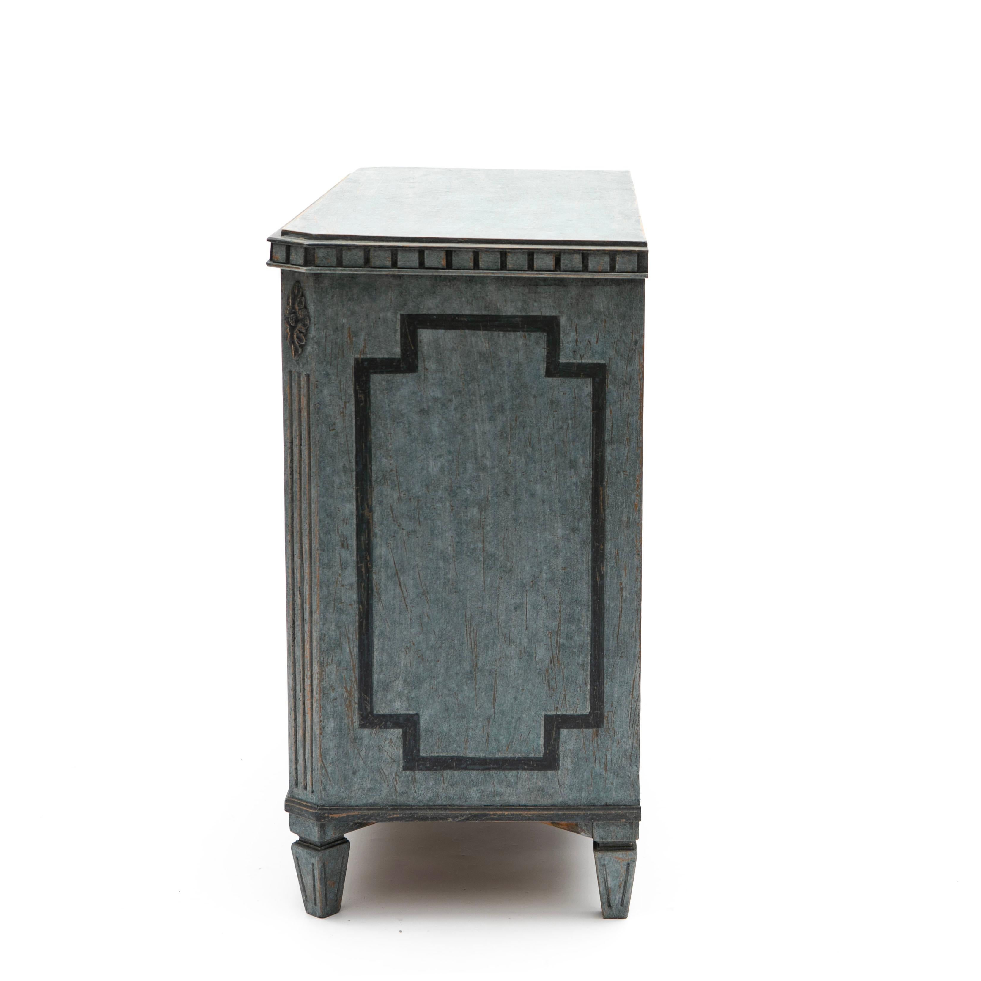 Decorative Gustavian-style chest of drawers in an elegant blue tone.
The front features three drawers adorned with a geometric pattern in a darker gray-blue shade.
The top drawer is narrow, followed by two larger drawers, all fitted with patinated