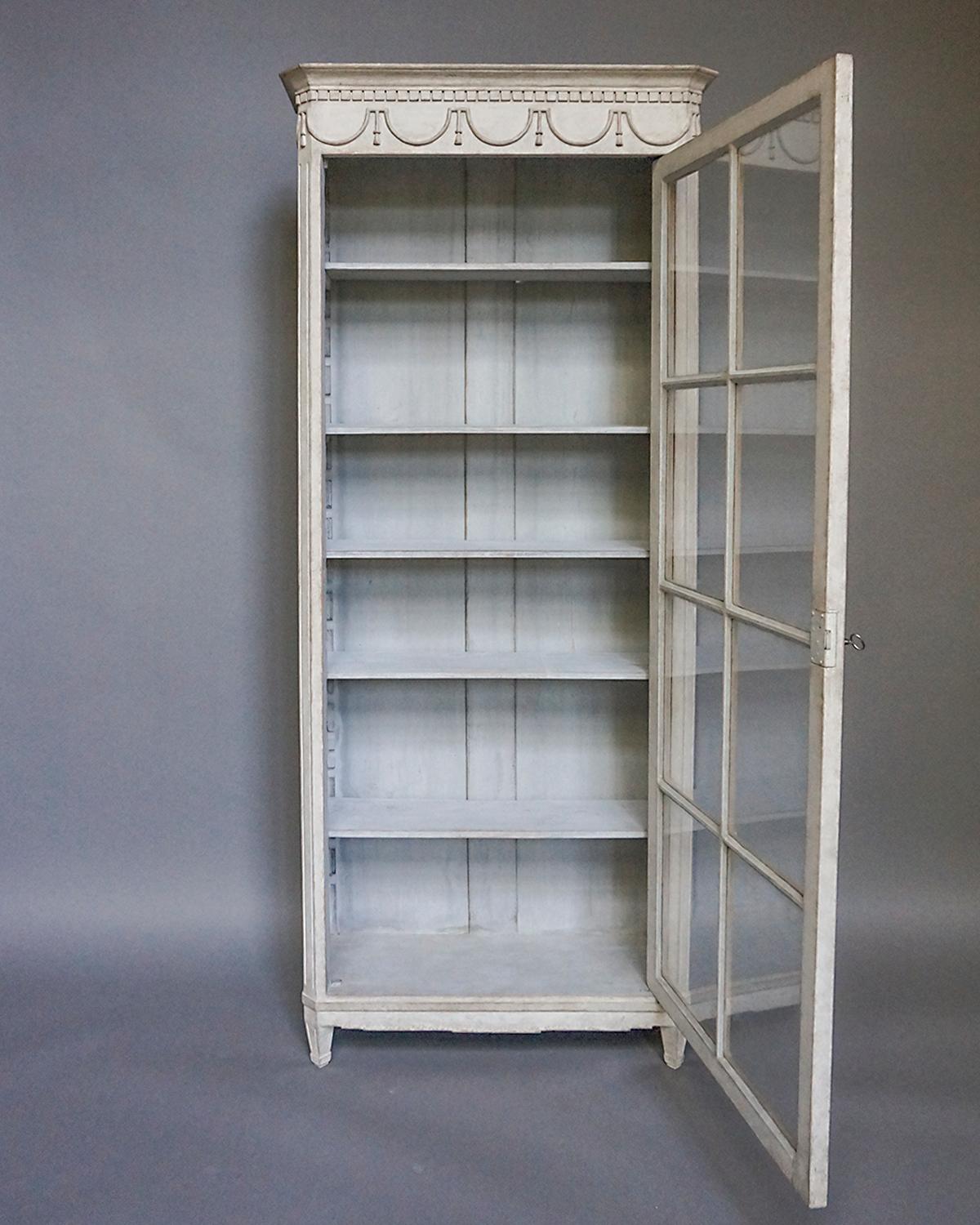 Glass-fronted cabinet or bookcase in the Gustavian style, Sweden circa 1880, with 8 individual panes. Simple molded cornice with dentil detail over a frieze of stylized swags and tassels. Canted corners with reeding over tapering square legs. Inside