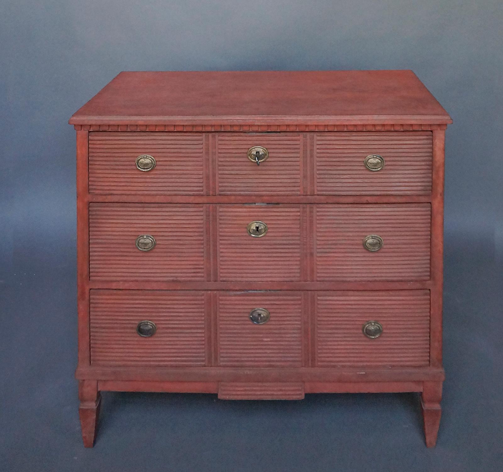 Three drawer chest in the Gustavian Style, Sweden, circa 1910, in a rich red ochre paint. The drawer fronts have horizontal reeding divided into three panels, with brass pulls and escutcheons. Dentil molding at the top, shaped apron, and tapering