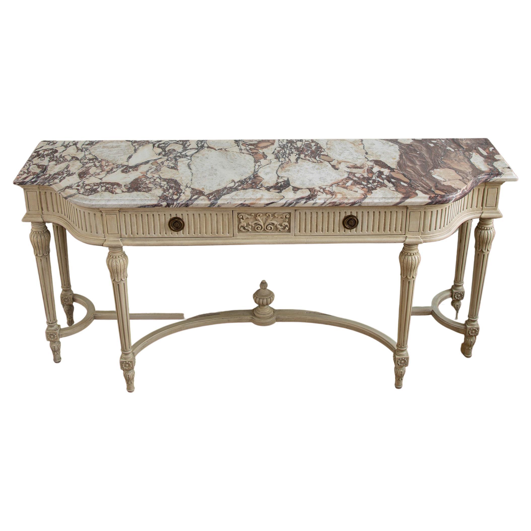 A hand carved/hand finished console in the Gustavian Style featuring Louis XVI detailing with two drawers in a lightly aged, antique white, patina. A further striking feature of this piece is the French Breche Violette marble top, beveled to shape.