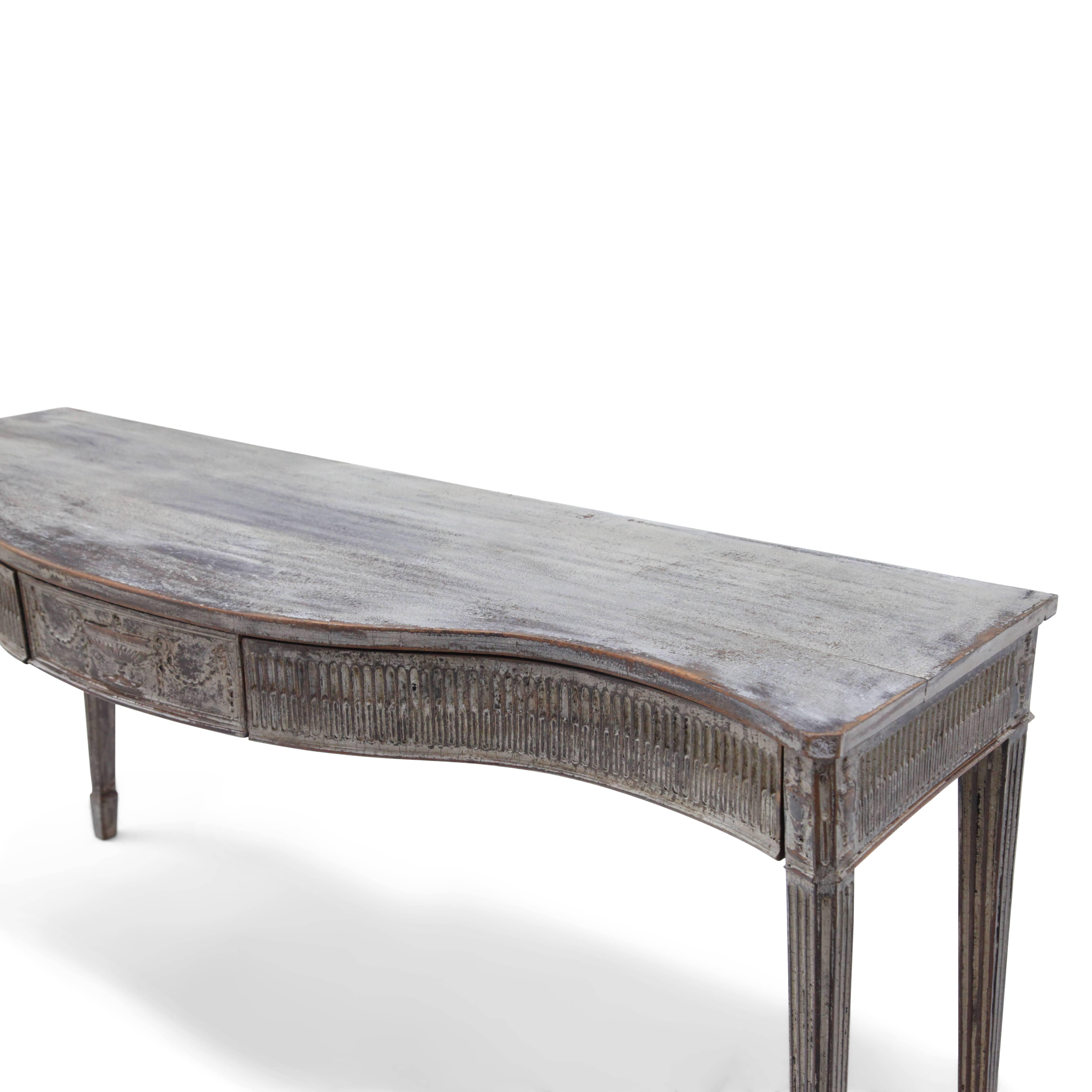 Two-drawered, Gustavian Style console table with a serpentine front on tall fluted legs. The apron is fluted as well and shows reliefs of amphorae in the centre and on the corners.