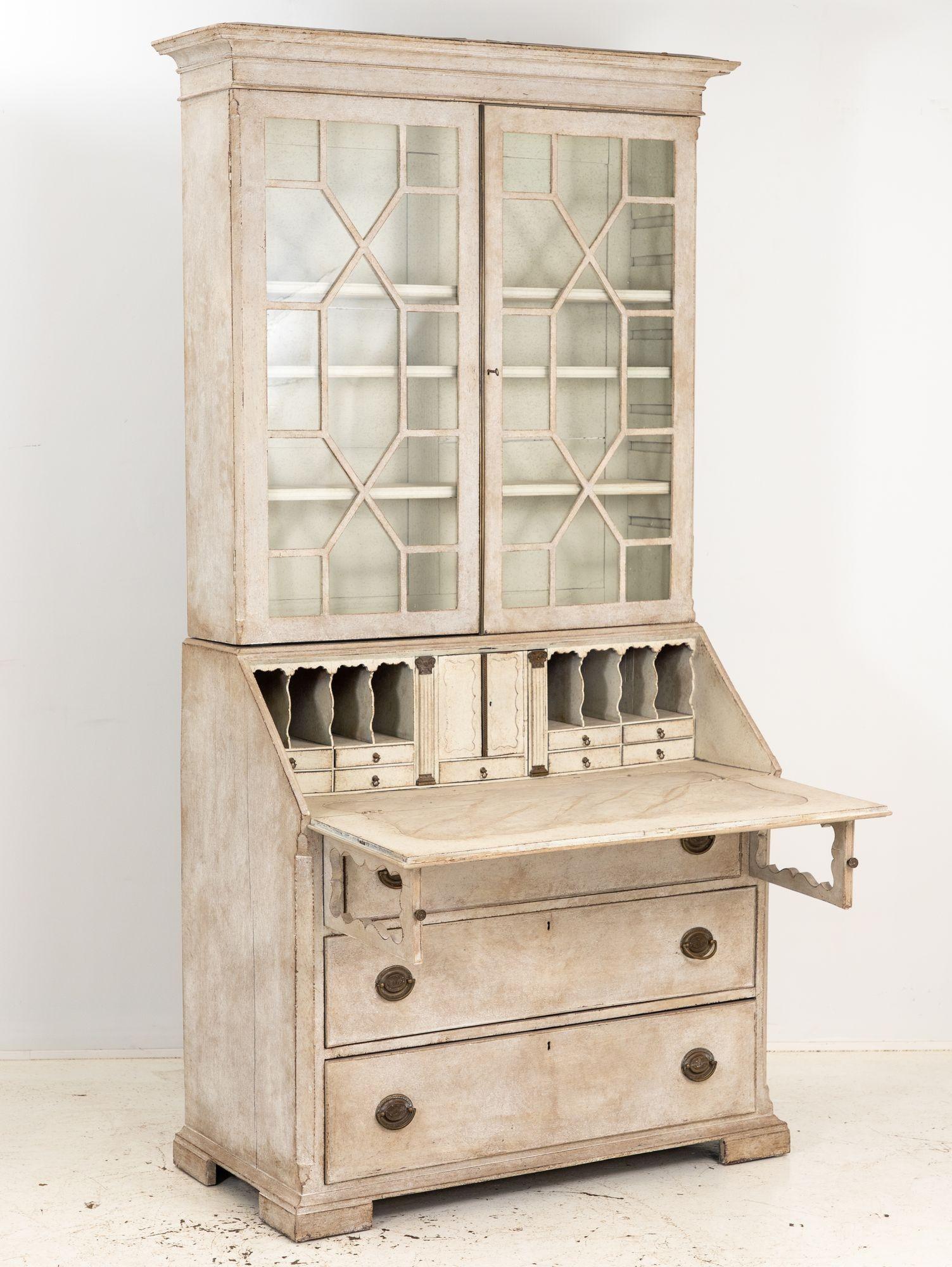 This late 19th-century Gustavian style drop-front secretary bookshelf is a true embodiment of timeless sophistication. Adorned with a later gray paint that enhances its antique allure, it retains its original glass with delicate grid-patterned