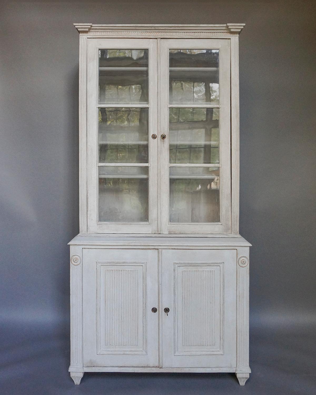 Two part cabinet in the Gustavian style, Sweden circa 1860. The upper section has a simple cornice with dentil molding and reeded corner posts. The double glazed doors open onto four fixed shelves. The lower section has a single shelf behind a pair