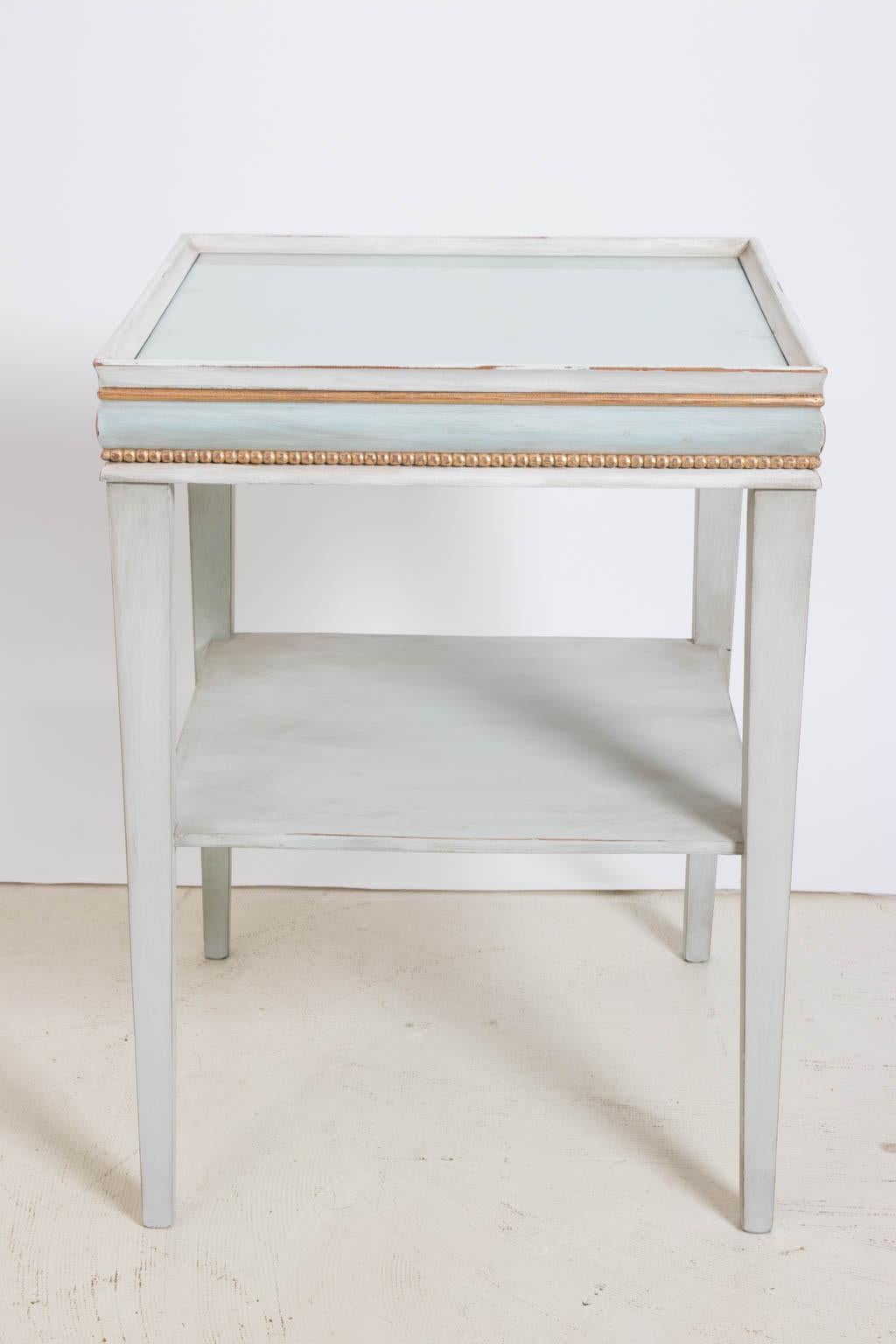 Painted square side table in the Gustavian style with glass insert top and bottom shelf, circa 1950s. The piece features beaded giltwood trim along with decorative molding in subtle painted shades of blue. Please note of wear consistent with age