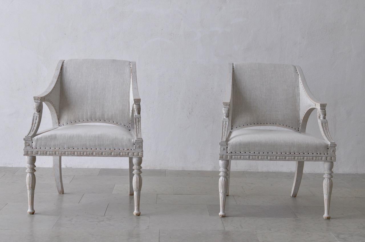 A special pair of painted, Gustavian style armchairs featuring sphinx heads on the arms and scroll backs. There is beautiful, carved leaf and floral motifs around the seat frame. These chairs have been newly upholstered in linen with nailhead