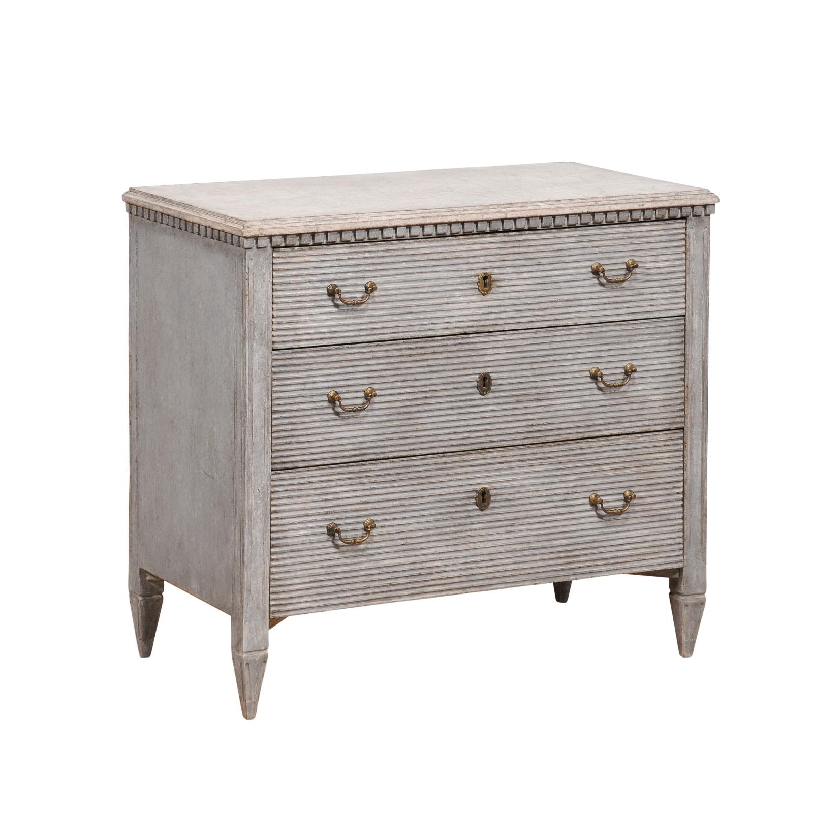 A Swedish Gustavian style chest from the 19th century with gray painted finish, three drawers, carved dentil molding and reeded accents. This Swedish Gustavian style chest, hailing from the 19th century, exudes timeless charm with its sophisticated