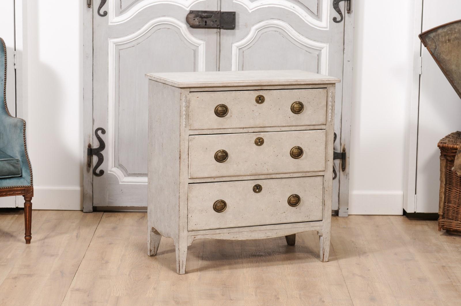 A Swedish Gustavian style light grey painted chest from the 20th century with three drawers, floral carved side posts, valanced apron and tapered feet. Immerse your living space in the timeless allure of Scandinavian elegance with this 20th-century