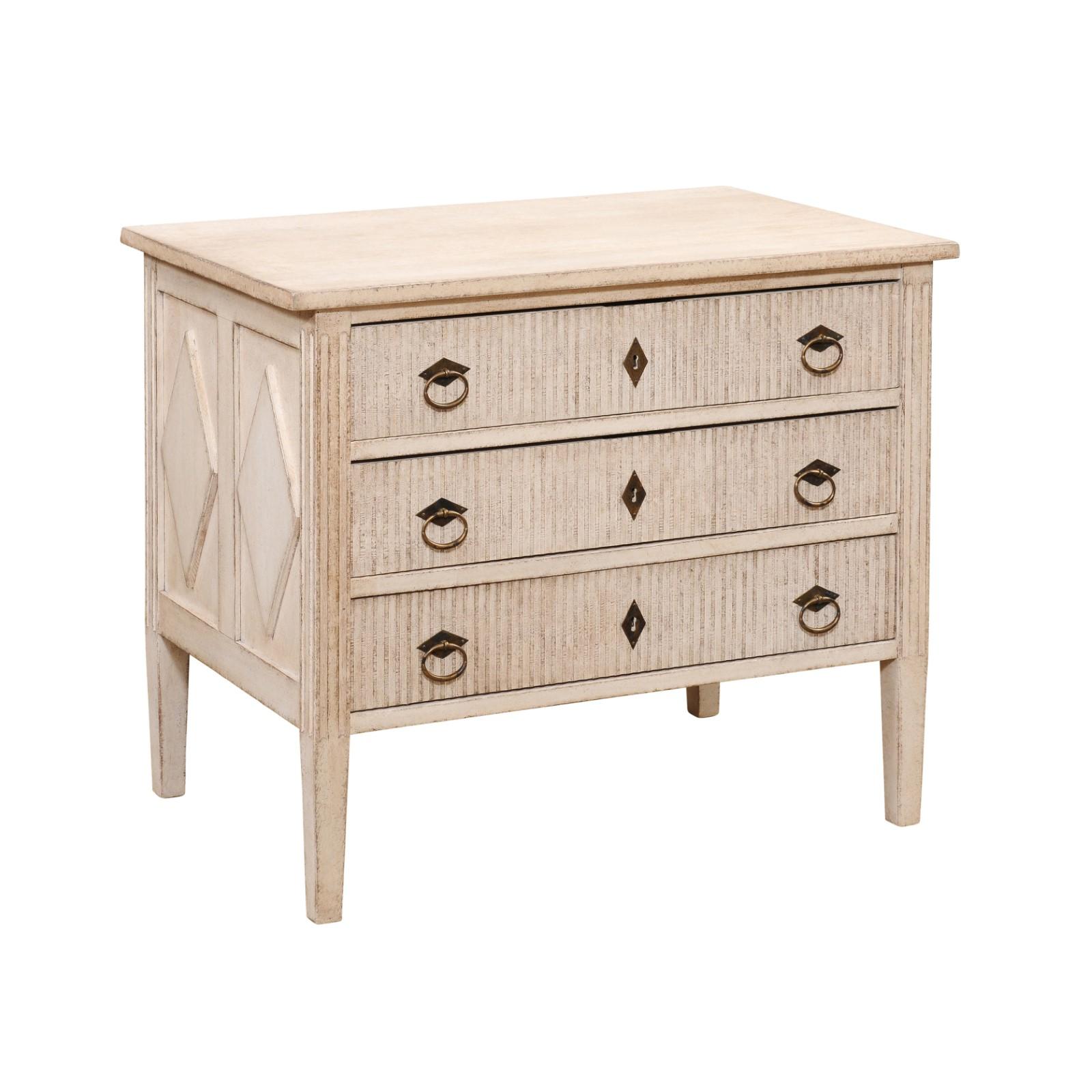 A Swedish Gustavian style three-drawer chest from the 19th century with creamy painted finish, carved reeded motifs on the drawers and diamonds on the side panels. Delve into the serene beauty of Swedish design with this 19th-century Gustavian style