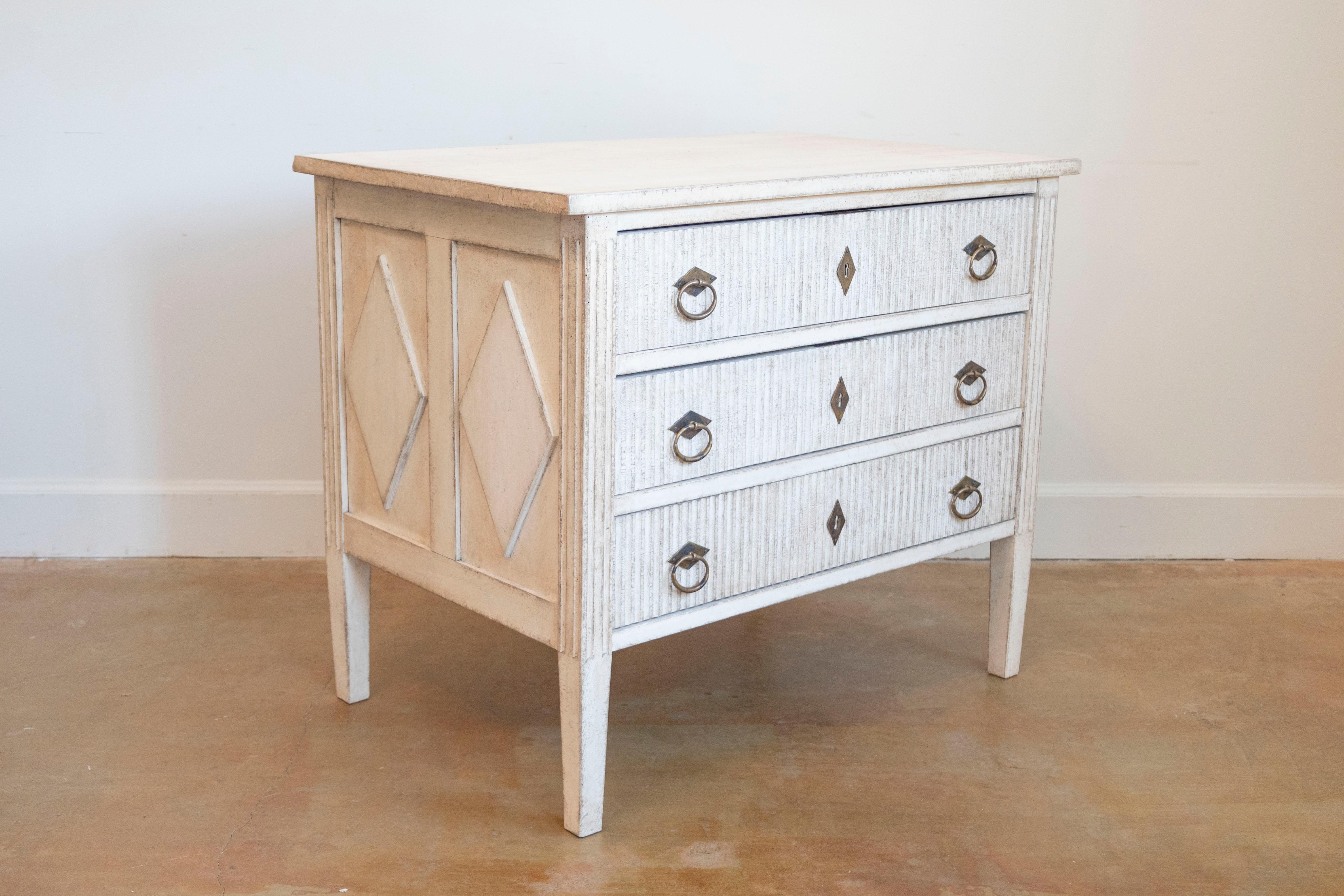 A Swedish Gustavian style three-drawer chest from the 19th century with creamy painted finish, carved reeded motifs on the drawers and diamonds on the side panels. Delve into the serene beauty of Swedish design with this 19th-century Gustavian style