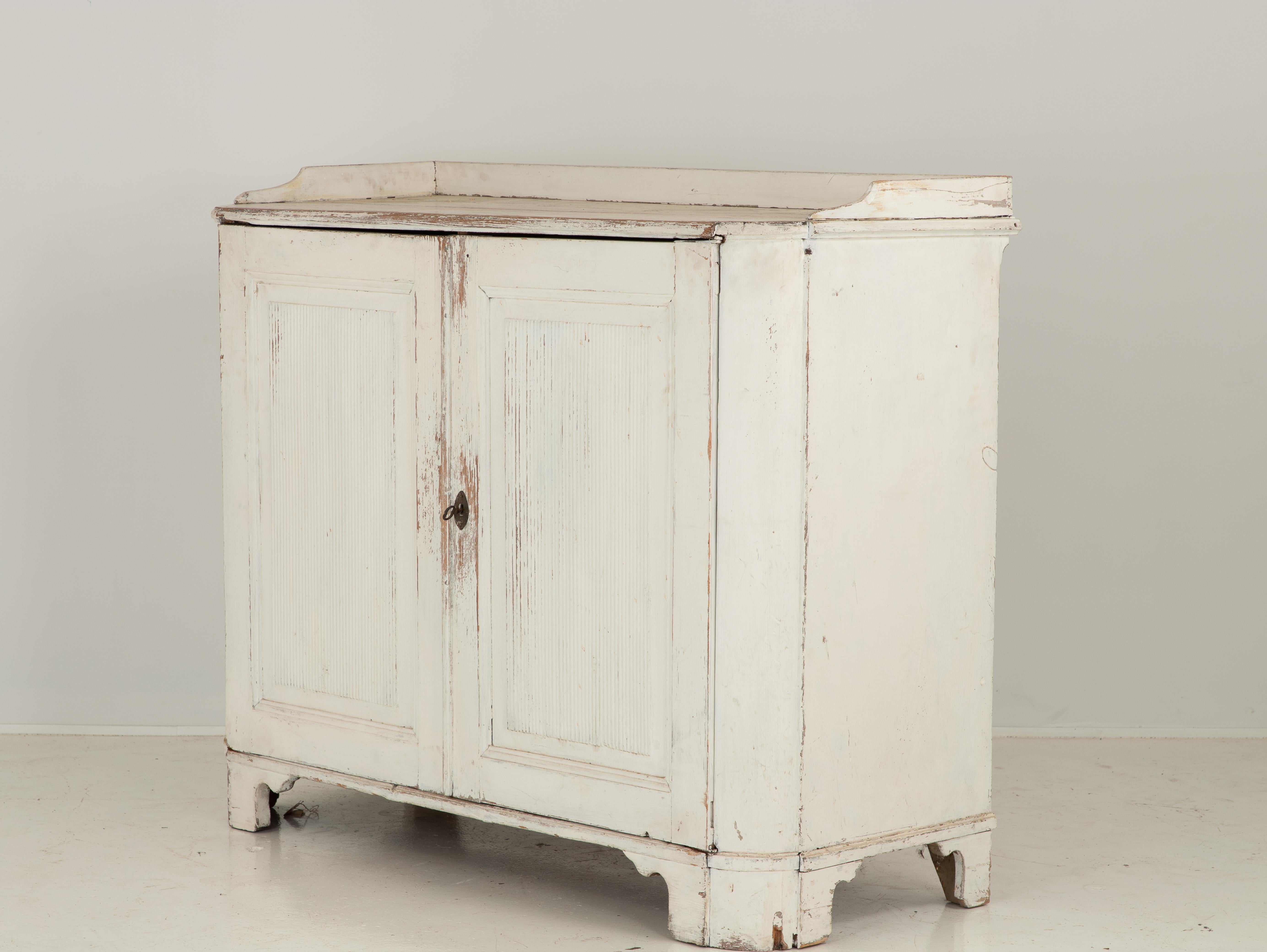 A white-painted Gustavian style buffet. Two reeded front doors with working lock and key open to reveal three fixed shelves and two drawers with dividers. The rounded corners terminate on bracket feet. The top is complete with a gallery. Beautifully