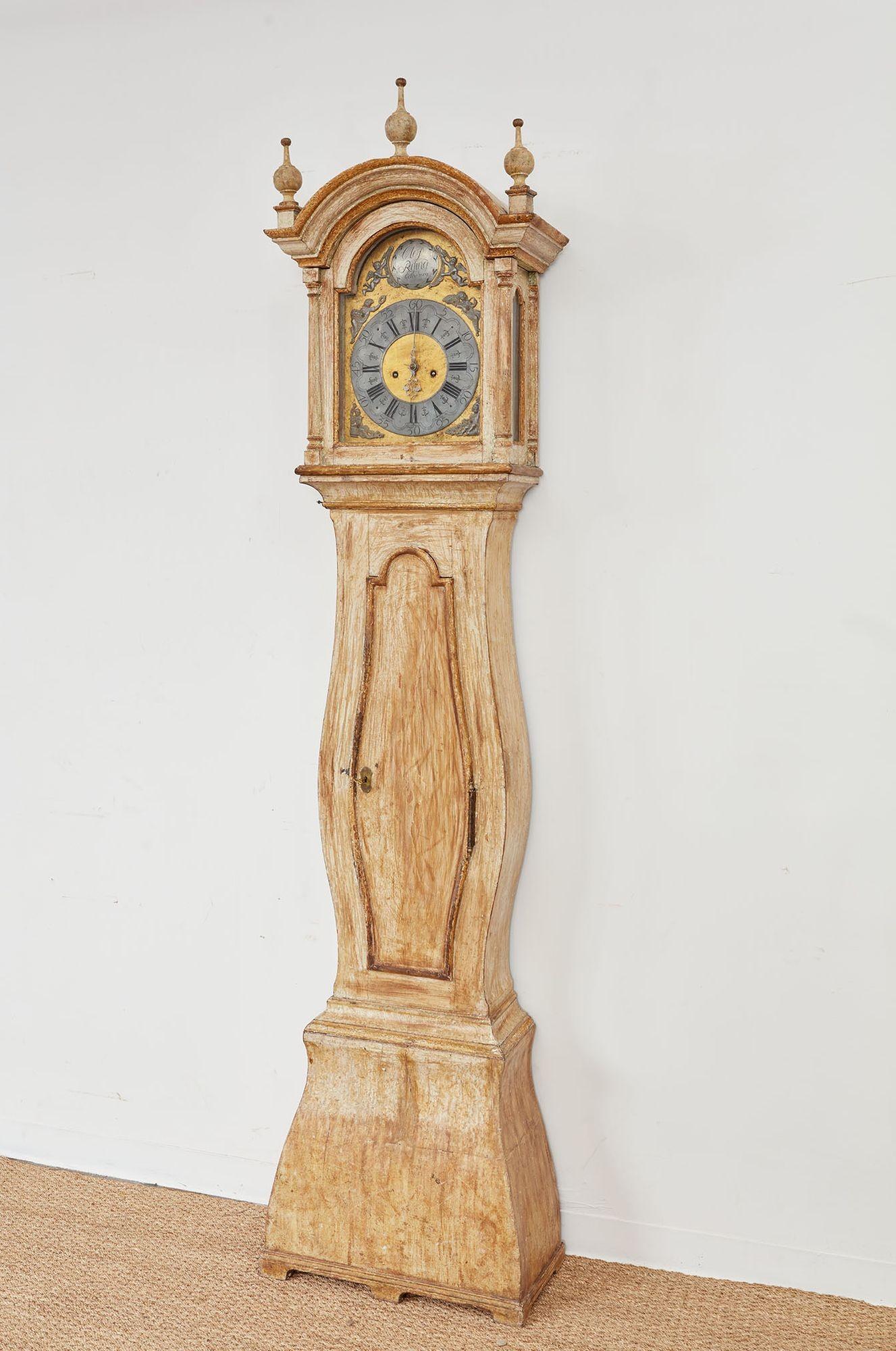 An elegant tall-case Gustavian clock having an imposing bonnet with an three-finial arched cornice on a sinuous bombe body with shaped door and narrow waist over a curved plinth, all in a lovely dry scraped lime white surface.  The desirable eight