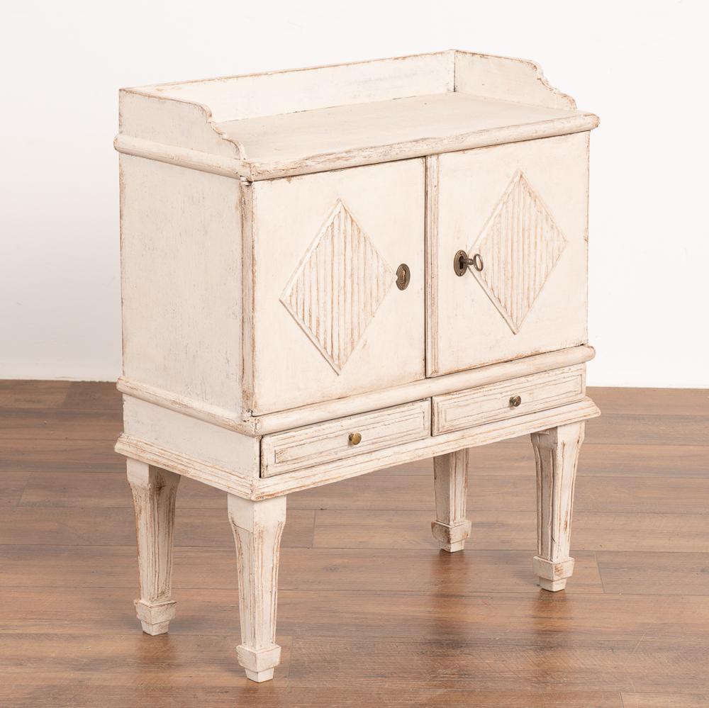 Small Swedish country white painted nightstand or side table with traditional decorative diamond motif on doors.
Two lower narrow drawers under the cabinet doors, resting on graceful tapered fluted legs. 
Newer professionally applied white painted