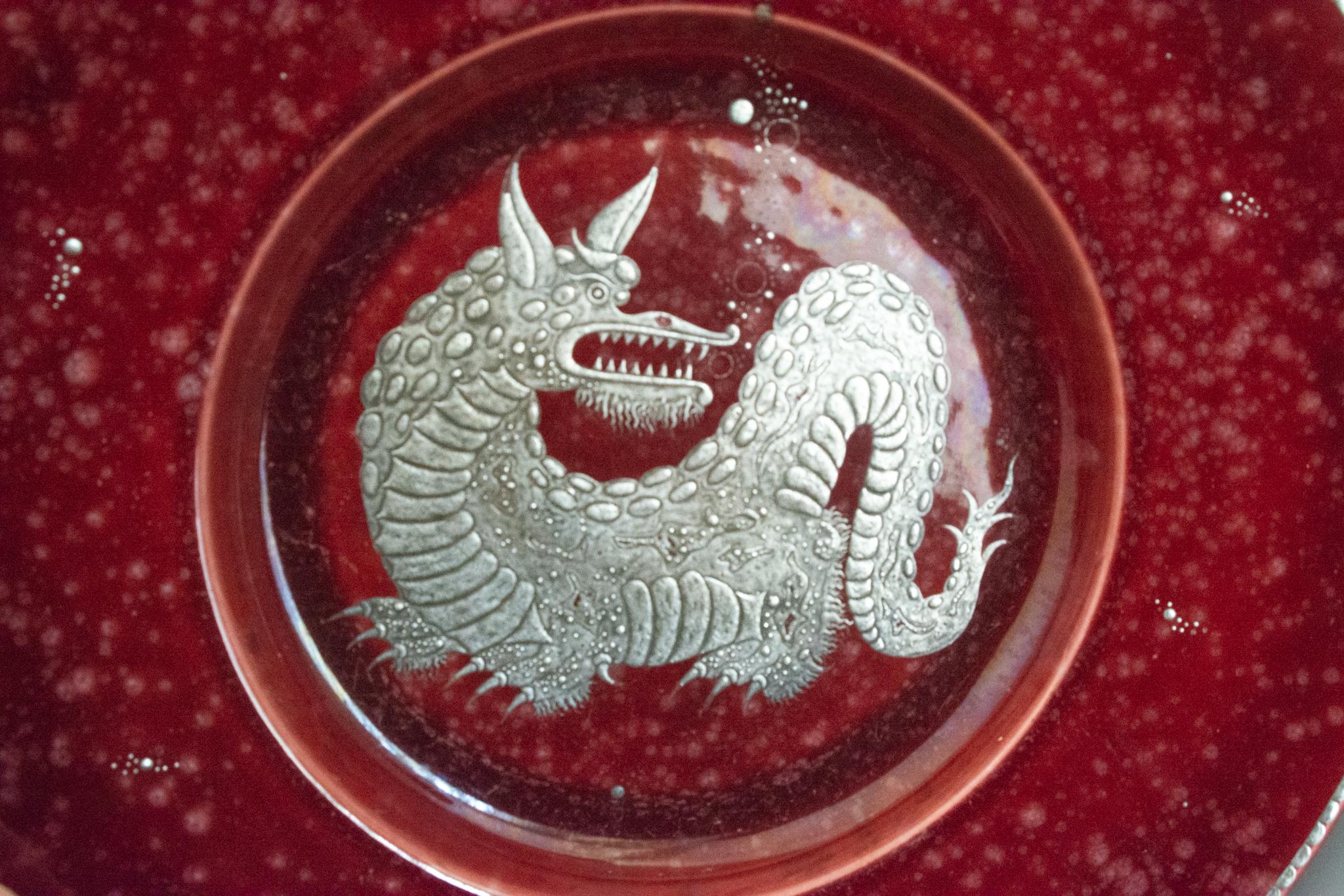 Un imponente caricatore raffigurante una creatura mitica in argento al centro, simile al Níðhöggr, un drago d'acqua su uno sfondo rosso intenso screziato. Le bolle d'aria argentate sono sparse sul piatto e ricordano il tema subacqueo che spesso