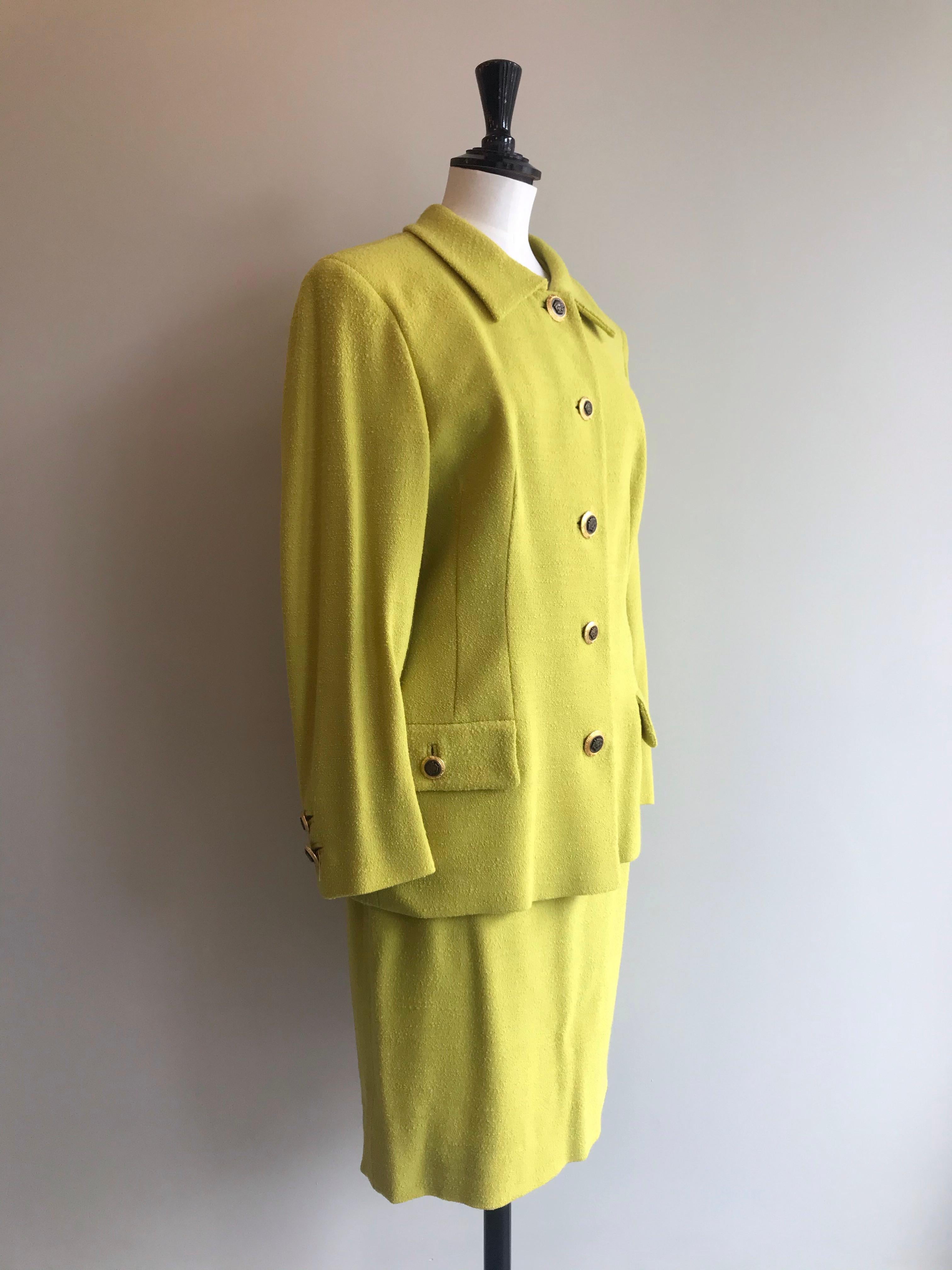 Chartreuse two piece wool skirt suit from Gianni Versace Versatile. Boxy blazer and straight cut knee length skirt. Statement Medusa adorned gold buttons in signature Versace style. Two closed off pockets at either side of the hip. Buttons on cuffs