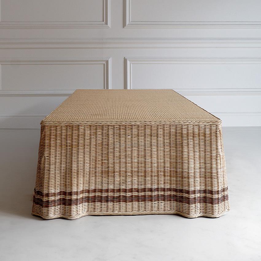 A coffee table handwoven in natural rattan, featuring a draped base mimicking the look of a cloth-covered table, crafted entirely from woven rattan for a structured, sculptural silhouette. A subtle brown stripe near the base, adds visual interest