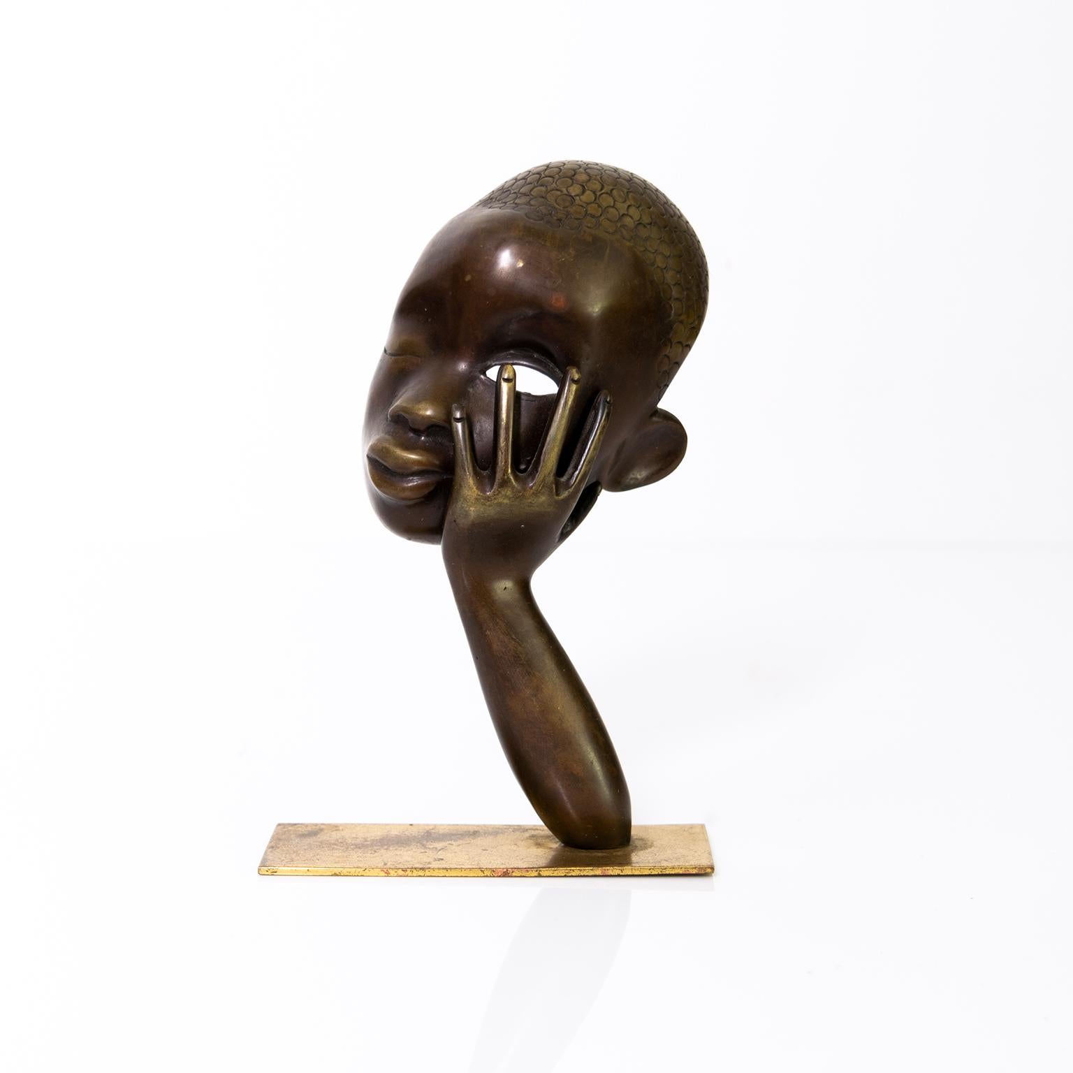 A bronze sculpture depicting a lovely female face mask being supported by one arm, produced by Hagenauer, Vienna Austria. 

Measures: Height: 6”, Width: 4”. Depth: 2.5”.