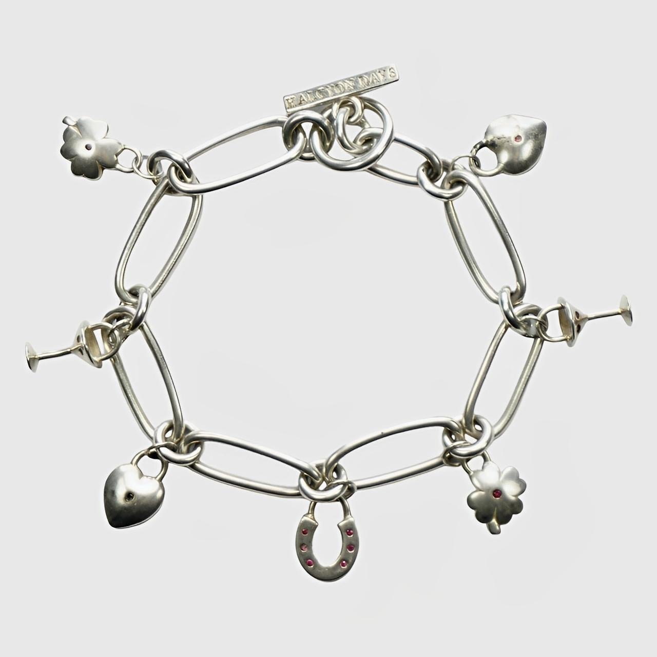 
Delightful Halcyon Days sterling silver link charm bracelet. There are seven charms: two hearts with red stones, two glasses, two four leaf clovers with red stones, and a horseshoe with red stones. 

The bracelet is stamped with a leopard's head,