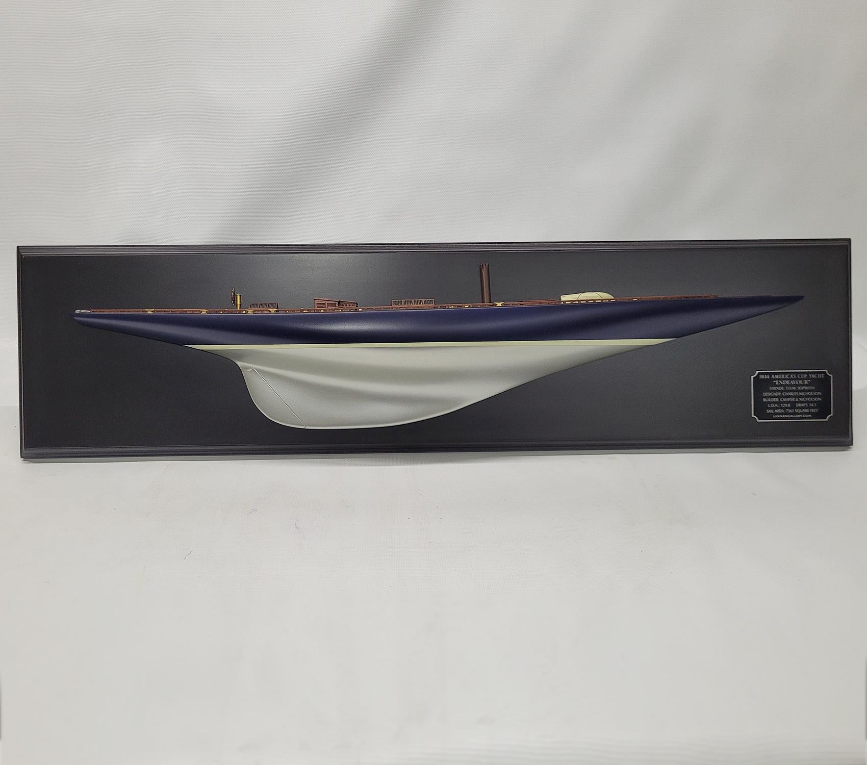Demi-modèle du Yacht Endeavor de la Coupe de l'America. Il s'agit d'une coque de 3 pieds montée sur un tableau noir. La coque est peinte en bleu sur fond argenté. Le pont en planches est équipé de lucarnes, d'un chemin d'accompagnement, d'un tangon,