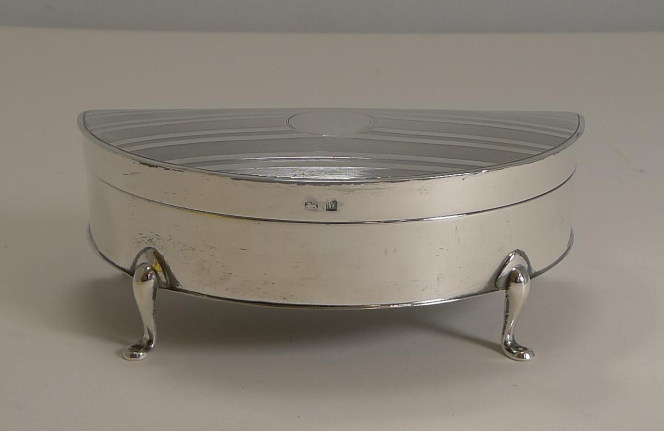A wonderful demilune or half-moon shaped jewellery box made from sterling silver standing on four elegant cabriole legs.

The top of the lid is engraved with a smart linear engine turned decoration with a central circular vacant cartouche.

The