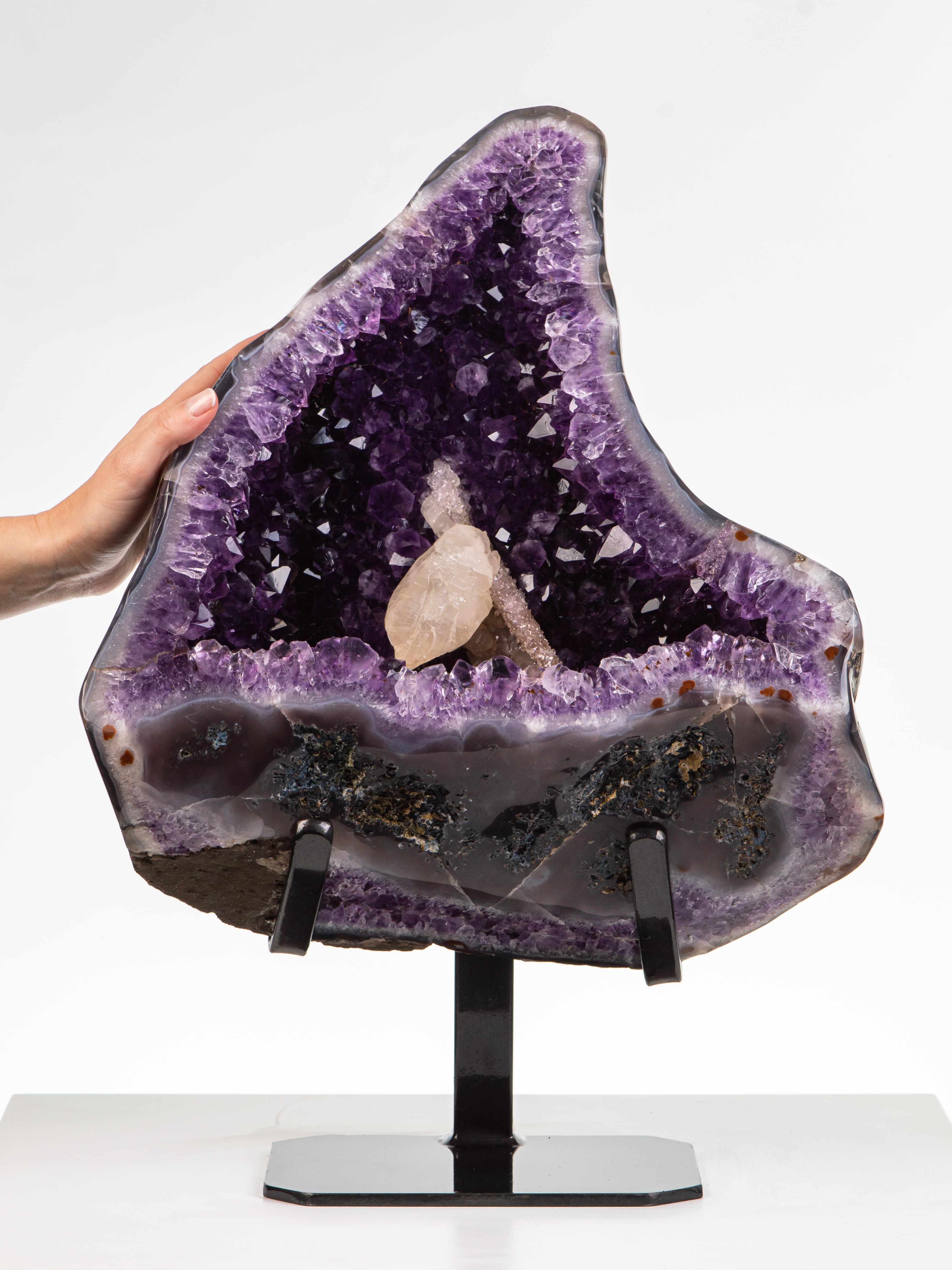 Eine halbpolierte Geode mit einem dicken agatierten Rand. Das Innere ist mit tiefvioletten Amethystkristallen und 2 schönen Kalzitformationen in der Mitte bedeckt, von denen eine mit einer Decke aus Drusus überzogen ist.

Dieses Stück wurde legal