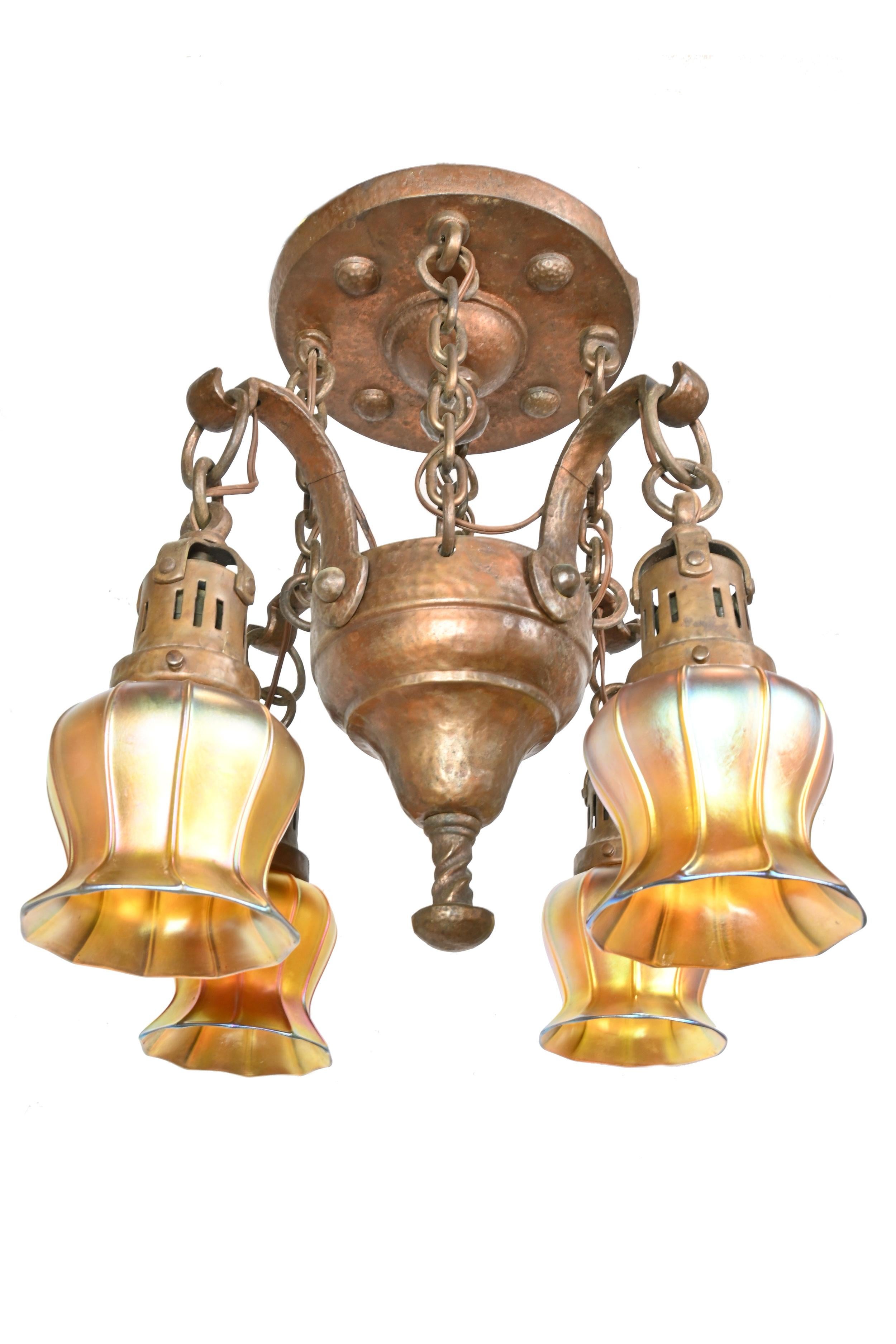 Beautiful finish on this hammered bronze Tudor fixture is almost as colorful as the stamped Quezal Aurene shades affixed to it. While the actual dimensions of the fixture are almost square this piece draws your eye vertically. 
Please note the
