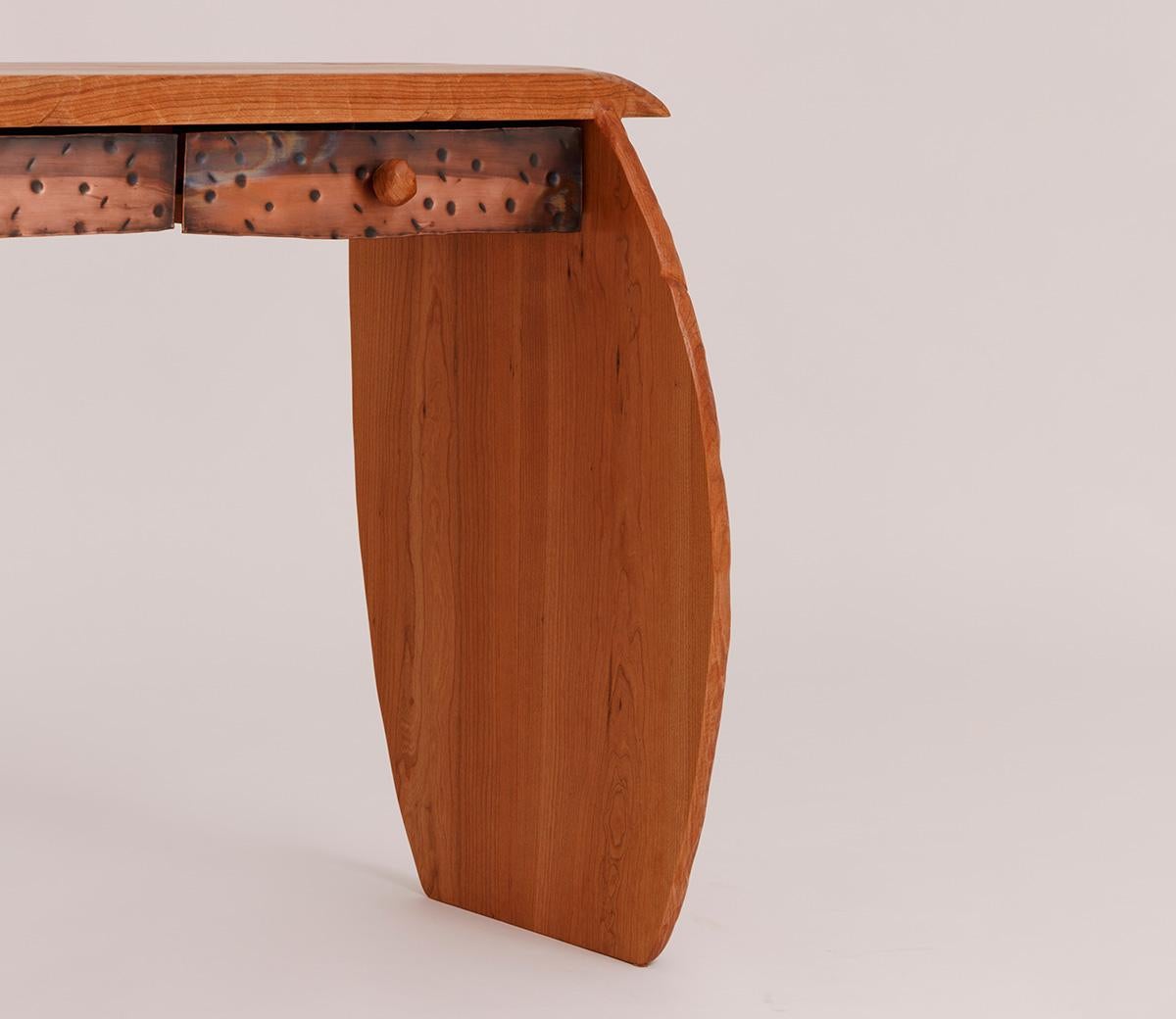 La consola Reno, tallada a mano en madera de cerezo macizo con frentes de cajón de cobre martillado, luce juguetones pomos de cerezo macizo. La pieza es totalmente personalizable.