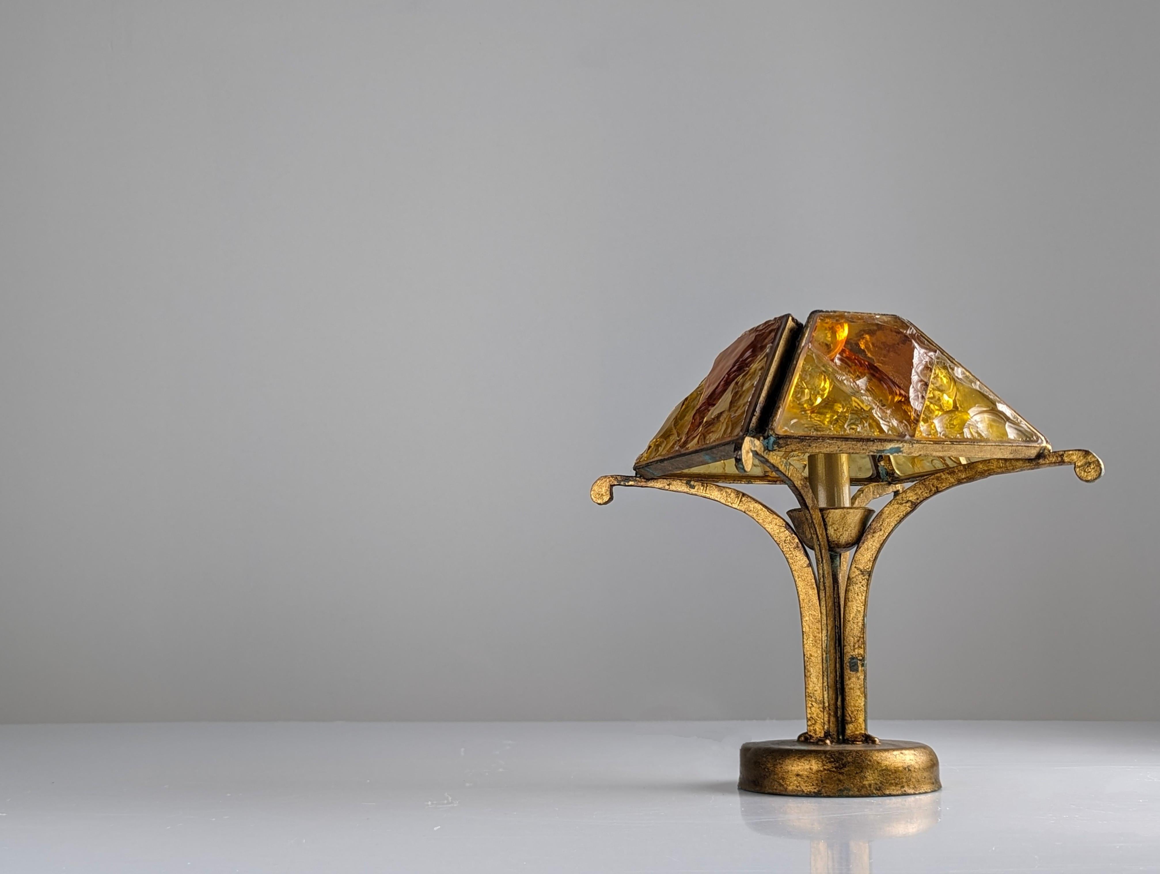 An extraordinary table lamp from the renowned Italian manufacturer Longobard, iconic of the Brutalist style of the 1970s. A beautiful piece made of wrought iron with a gilded finish, featuring thick hammered caramel-colored glass crystals that