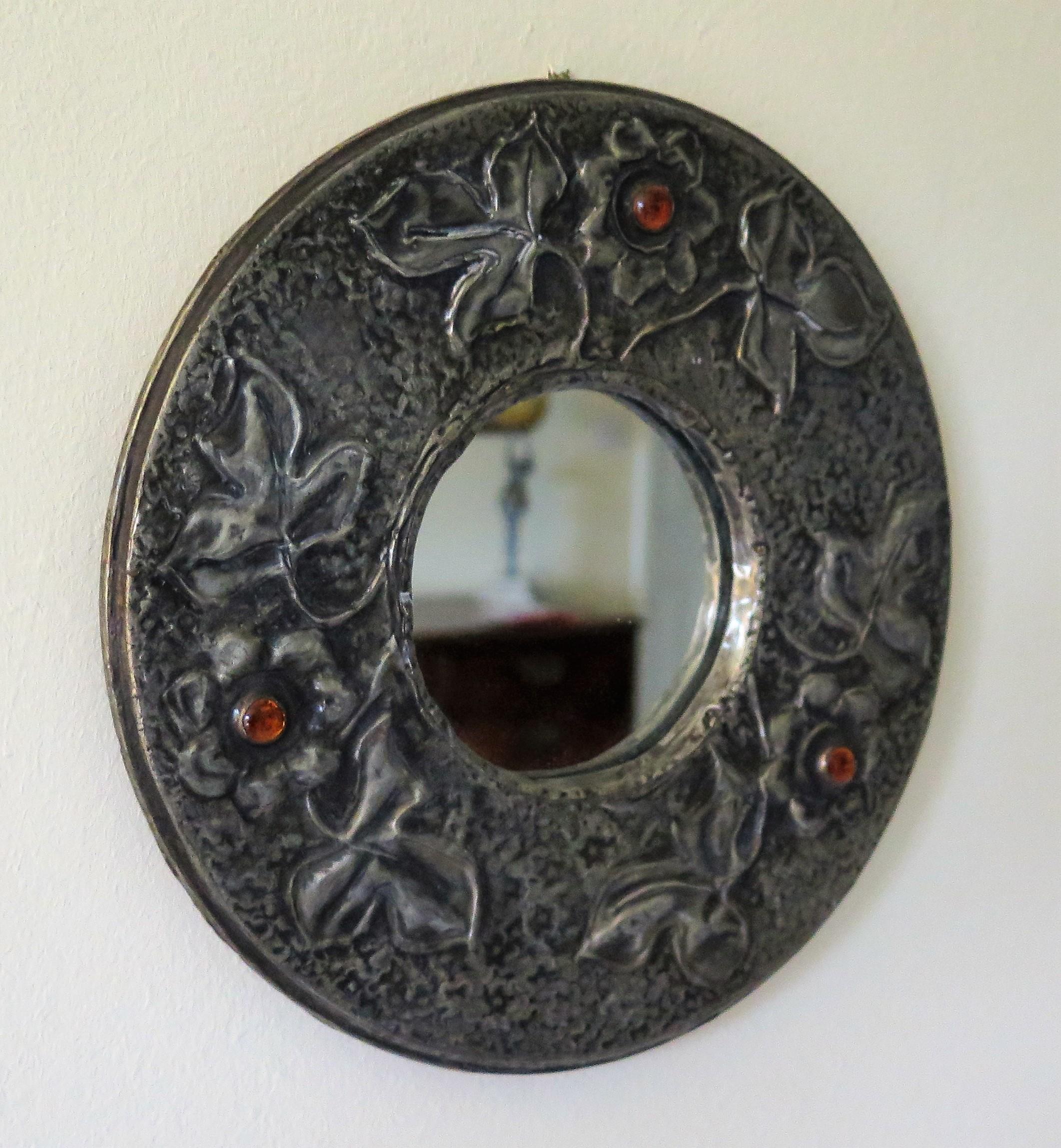 This is a very decorative English circular wall mirror of small diameter, made of hammered Pewter, with amber glass cabochons and dating to the English Arts and Crafts period, circa 1900

The mirror is circular with a central mirror glass with a