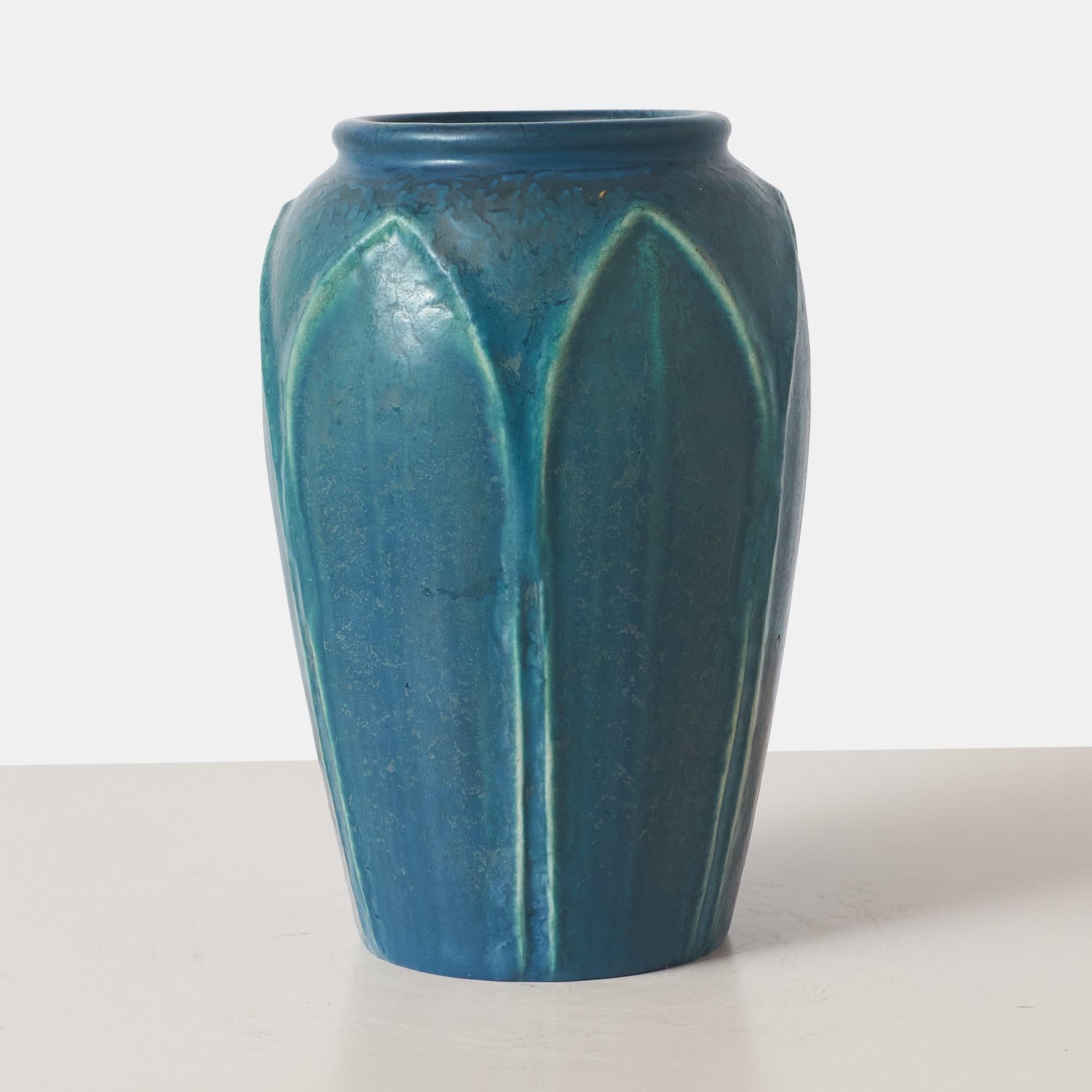 This Hampshire Pottery vase, designed by Cadmon Robertson, features five tall leaves. The vase is glazed in the signature two-tone peacock glaze that Hampshire Pottery is known for. The piece is marked Hampshire Pottery with a circled M logo as a
