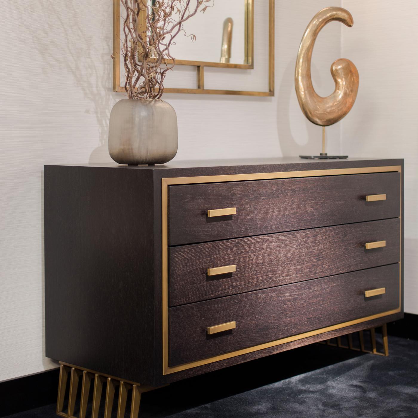 Standing on two openwork metal legs with a brass finish, this wooden dresser exudes restrained elegance and is be a perfect addition to a wide variety of refined interiors. Composed of three drawers with brass-finished metal handles, the structure
