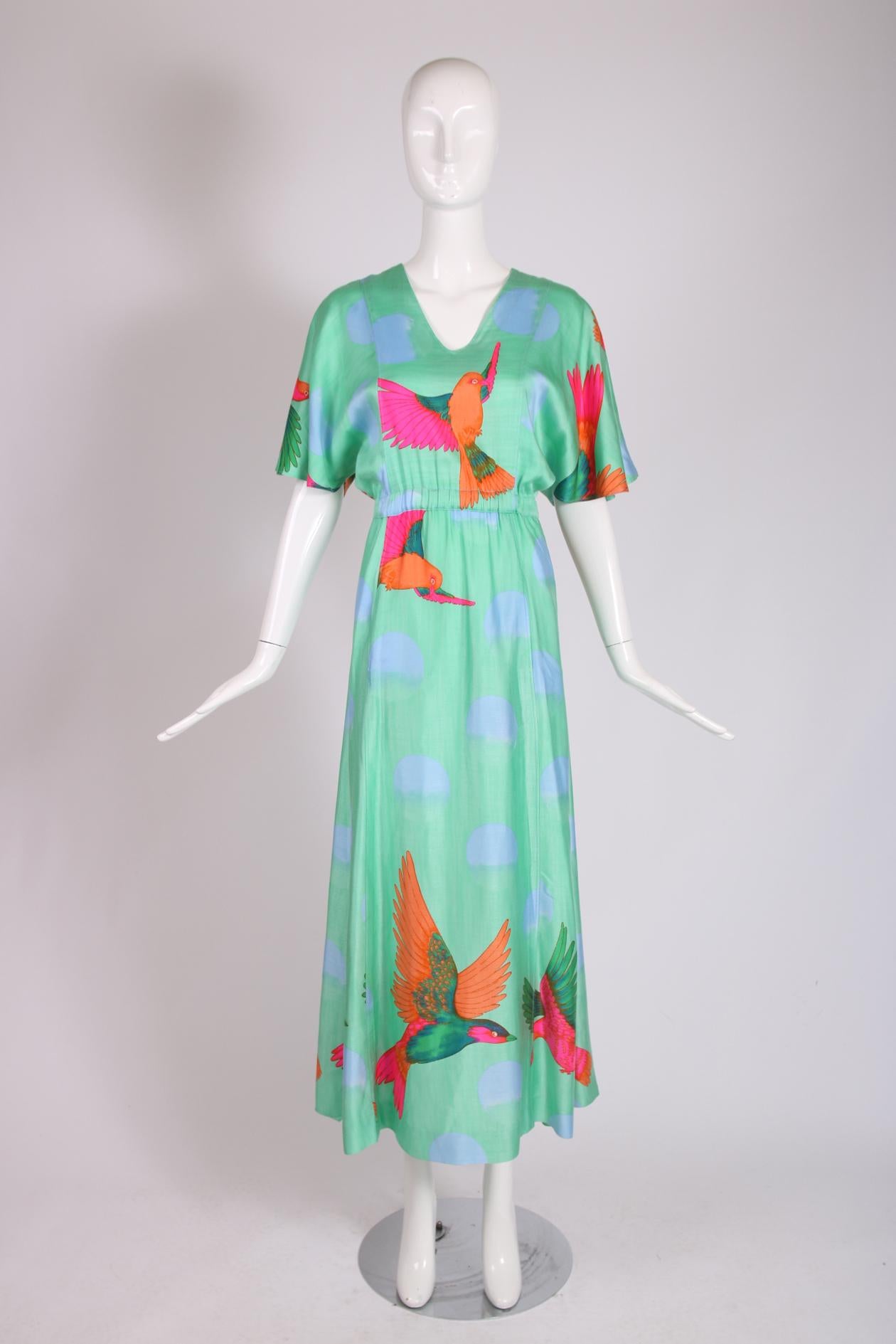 1970's Hanae Mori green flutter sleeve maxi dress with a v-neckline, cinched elastic waistline, two hidden frontal pockets and a print that features an exotic pink, orange and green bird set against a backdrop of repeating 3/4. purple moons. There