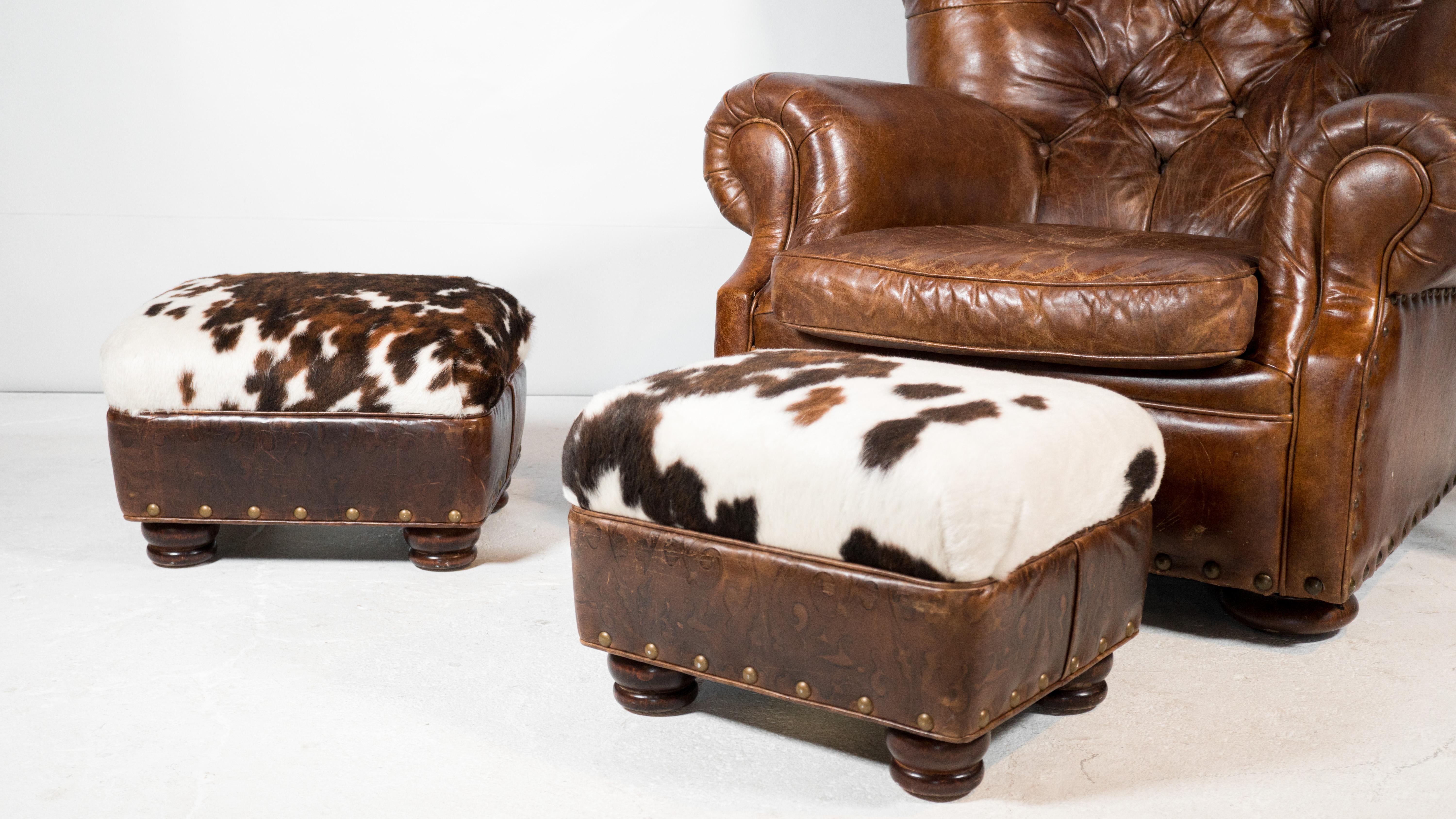 Hancock and Moore cowhide ottomans with brass nailhead trim. Beautiful cowhide pattern and opposing western floral brown leather siding. Brass nailhead trim and round wooden legs. Rustic cowboy design with modern aesthetics. Manufacturer label