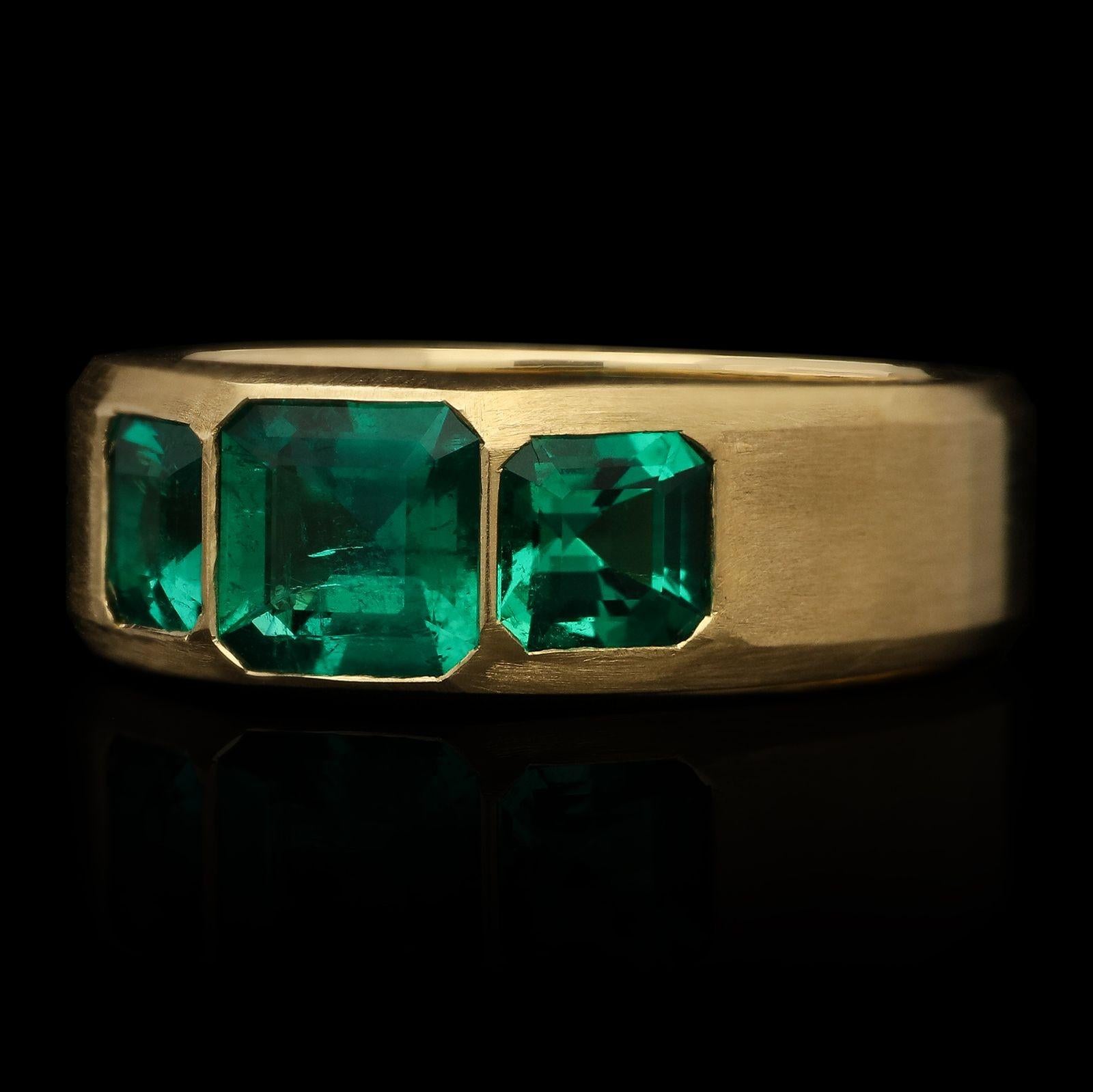 A striking emerald and gold three stone ring by Hancocks, centred with a Colombian emerald-cut emerald weighing 0.70ct set flush into the gold between two further Colombian emeralds weighing 0.84cts combined, all set in an 18ct yellow gold flat