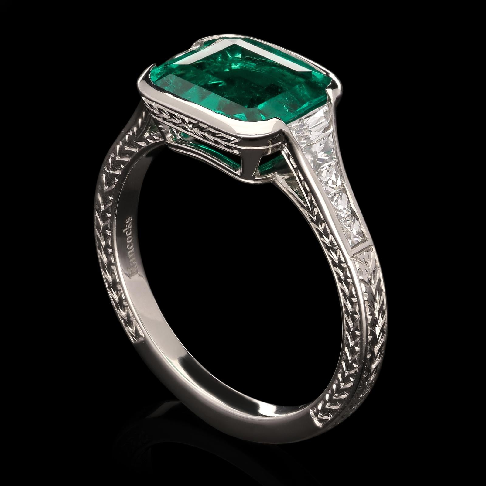 A beautiful emerald and diamond ring by Hancocks, centred with a cushion shaped untreated Colombian emerald weighing 2.42cts in partial rub over setting to elegantly tapering shoulders channel set with calibre French-cut diamonds, all in a finely