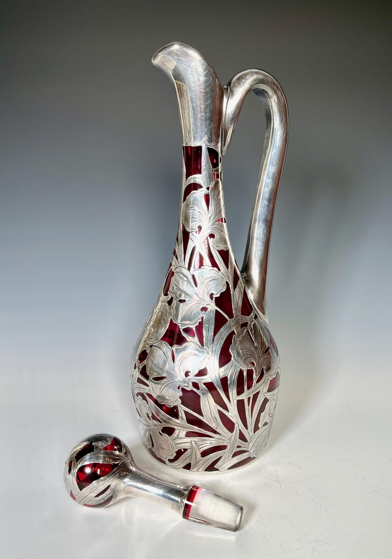 This tall hand blown cranberry red crystal decanter is an exquisite example of the masters of silver overlay decoration. The body is beautifully embellished with an allover Art Nouveau floral motif including the handle and stopper for an example of