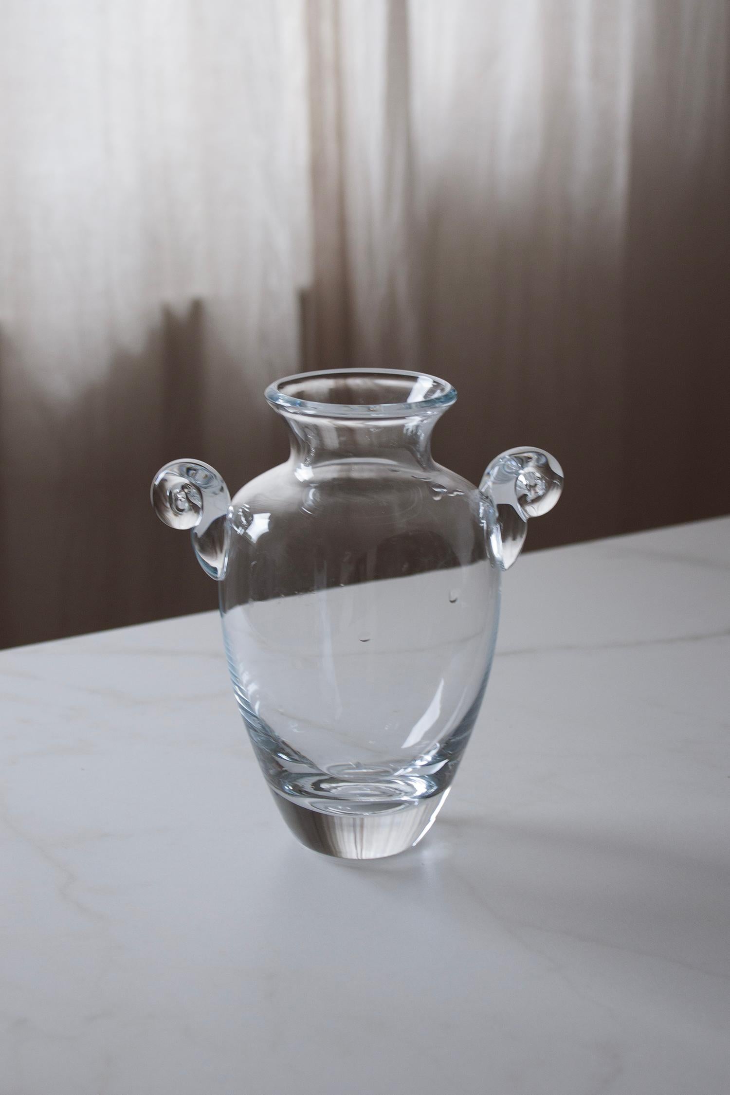 This elegant amphora-style vase is crafted from clear, hand-blown glass, featuring a classic baluster form with a flared rim and two distinctive scroll handles. Its restrained design highlights the purity and clarity of the glass, while the