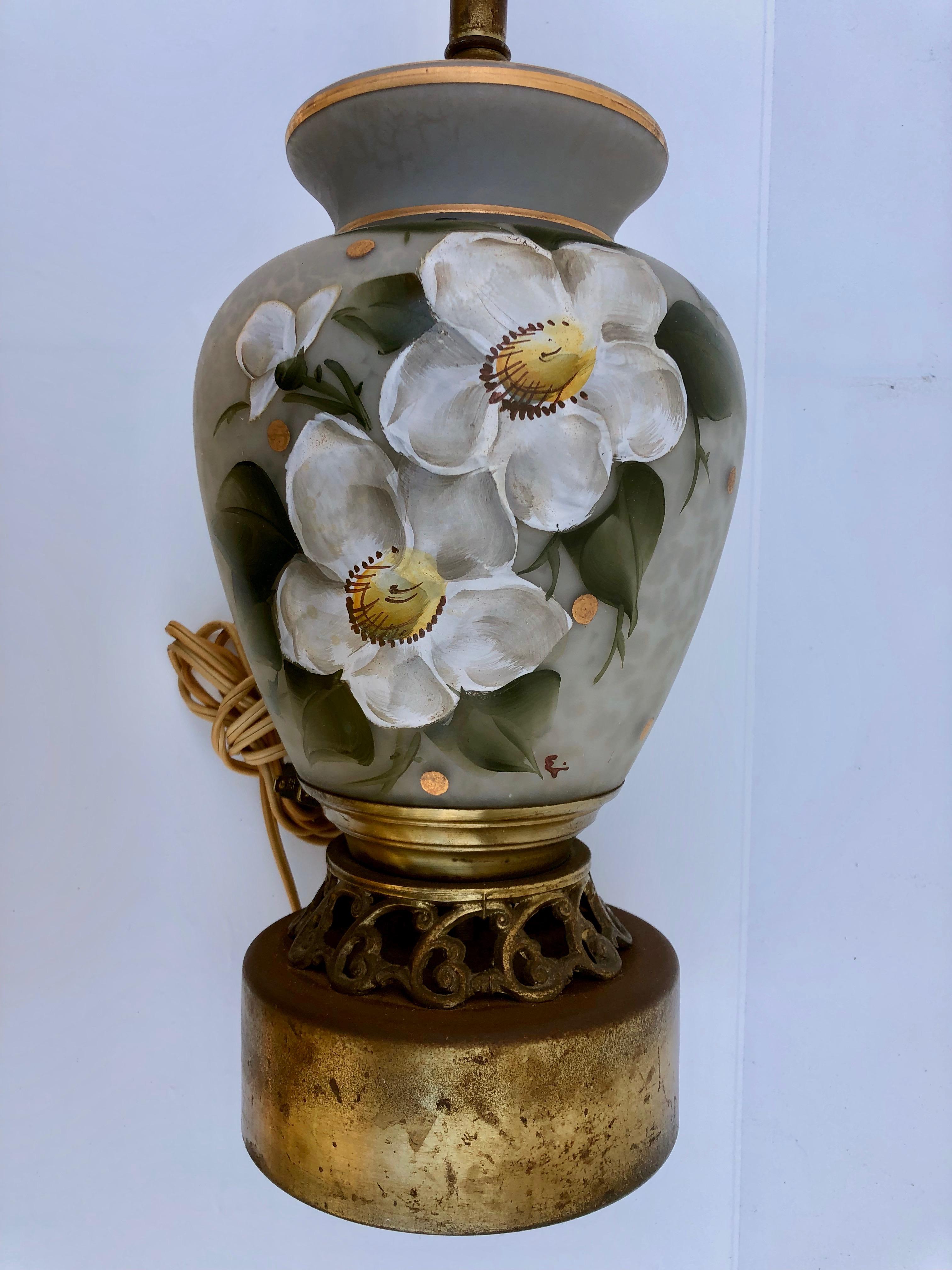 This is a stunning hand blown, hand painted glass lamp. It is a high quality hand painted lamp with an artist signature, as shown. The tone of the lamp is light grey, the two flowers are white with yellow and green leaves while there is gold trim