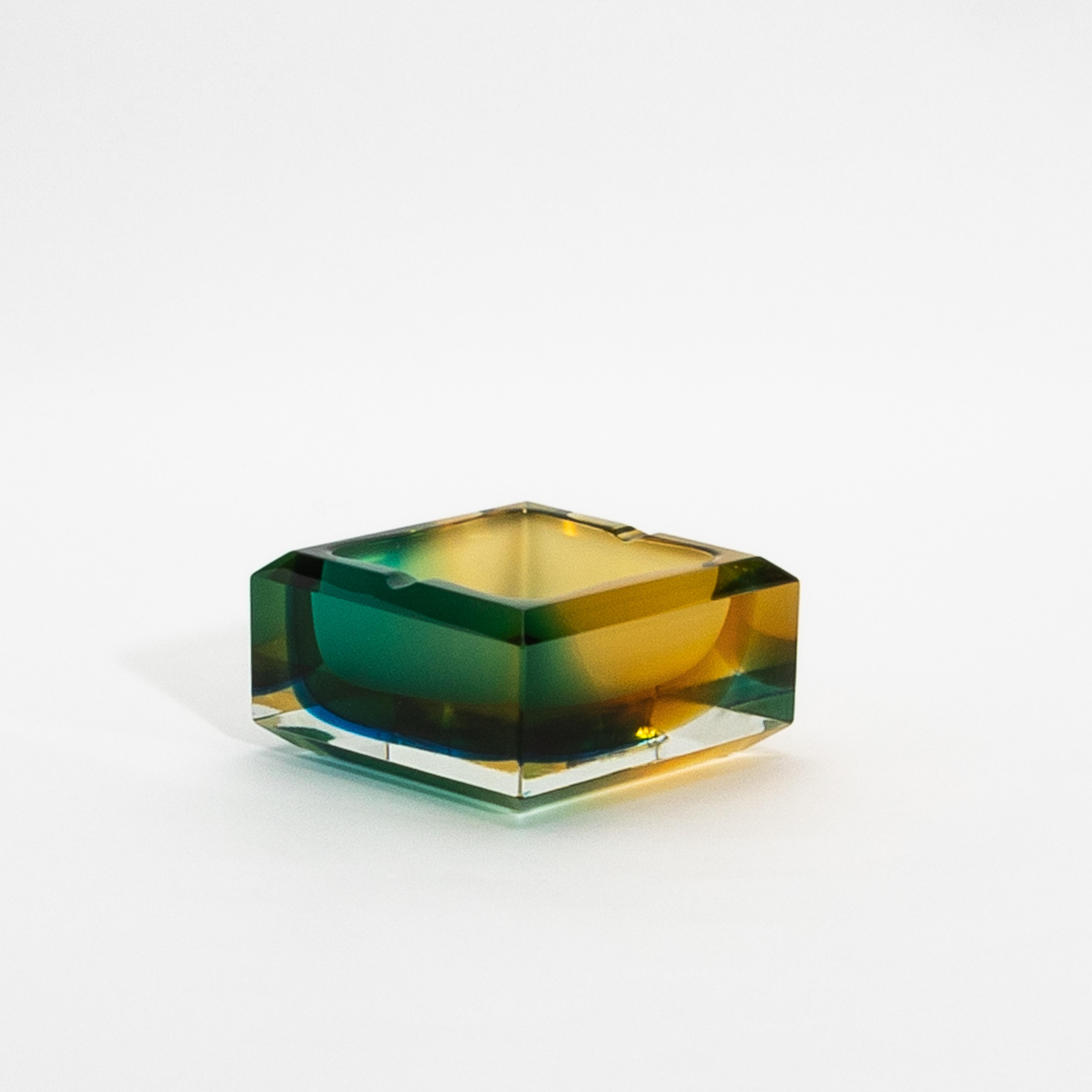 This exquisite hand-blown Murano glass ashtray embodies the unparalleled craftsmanship and luxury synonymous with Venetian design. The vibrant gradient of green, blue, and amber hues seamlessly melds together, creating an ethereal interplay of color