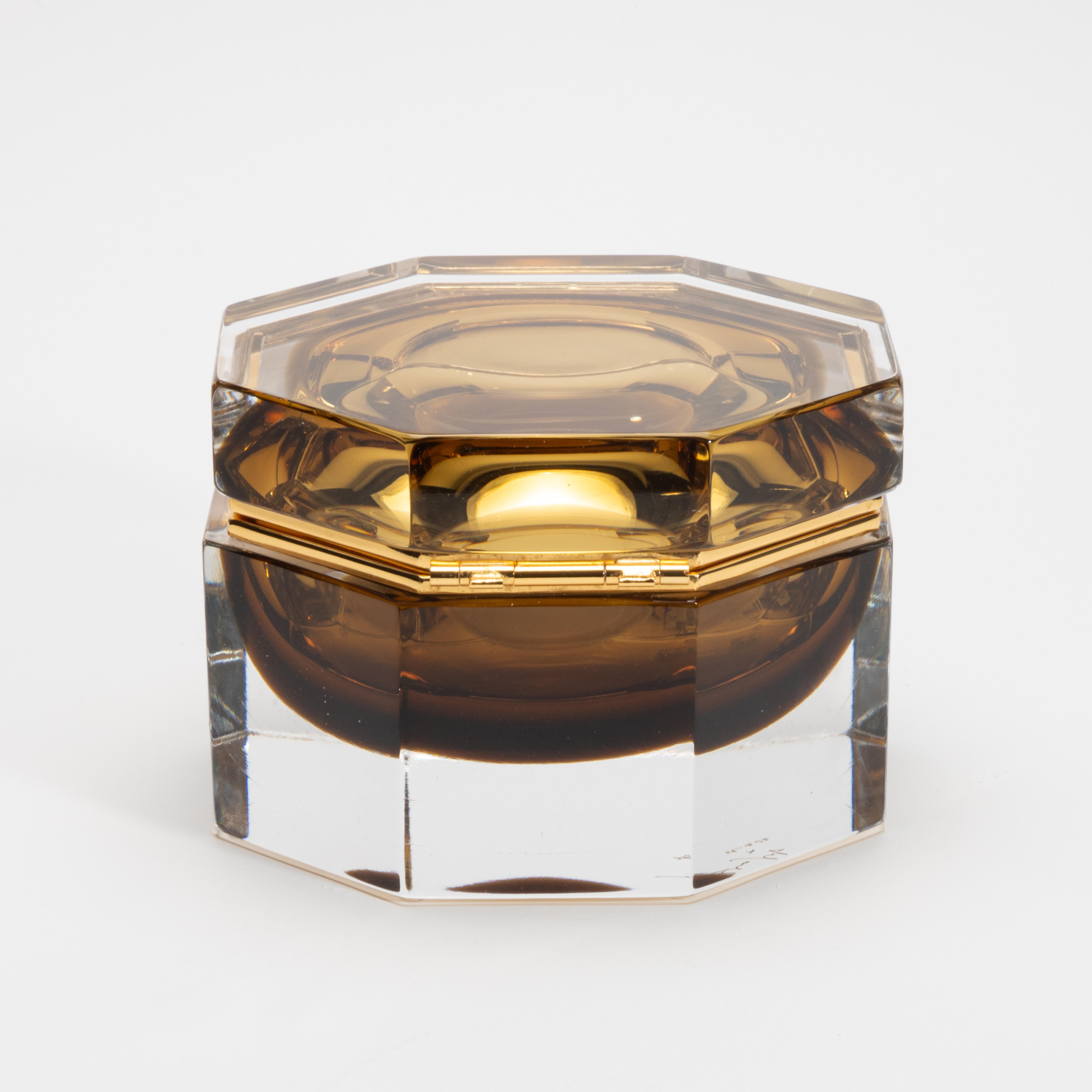 Diese exquisite achteckige Dose ist ein Fest der kühnen Geometrie und der handwerklichen Finesse, gefertigt aus mundgeblasenem Glas in einem satten Tabak-Ton. Die klare architektonische Silhouette umrahmt einen Innenraum, der sich mit skulpturaler