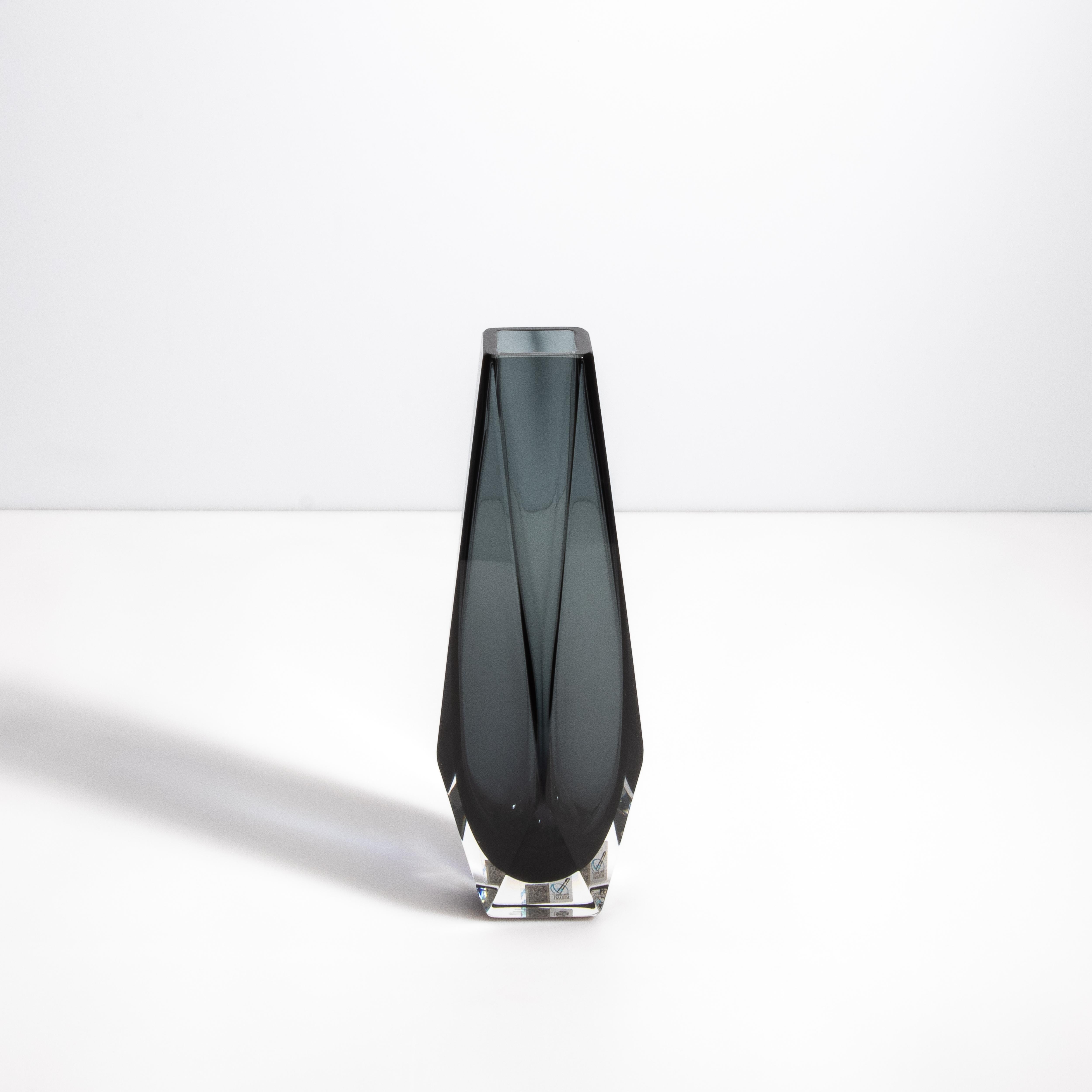 Questo vaso di Murano emana un fascino contemporaneo, fondendo la precisione geometrica con l'ispirazione organica di una goccia di pioggia. Realizzato da maestri vetrai, questo pezzo presenta una sorprendente tonalità di grigio intenso, incastonata