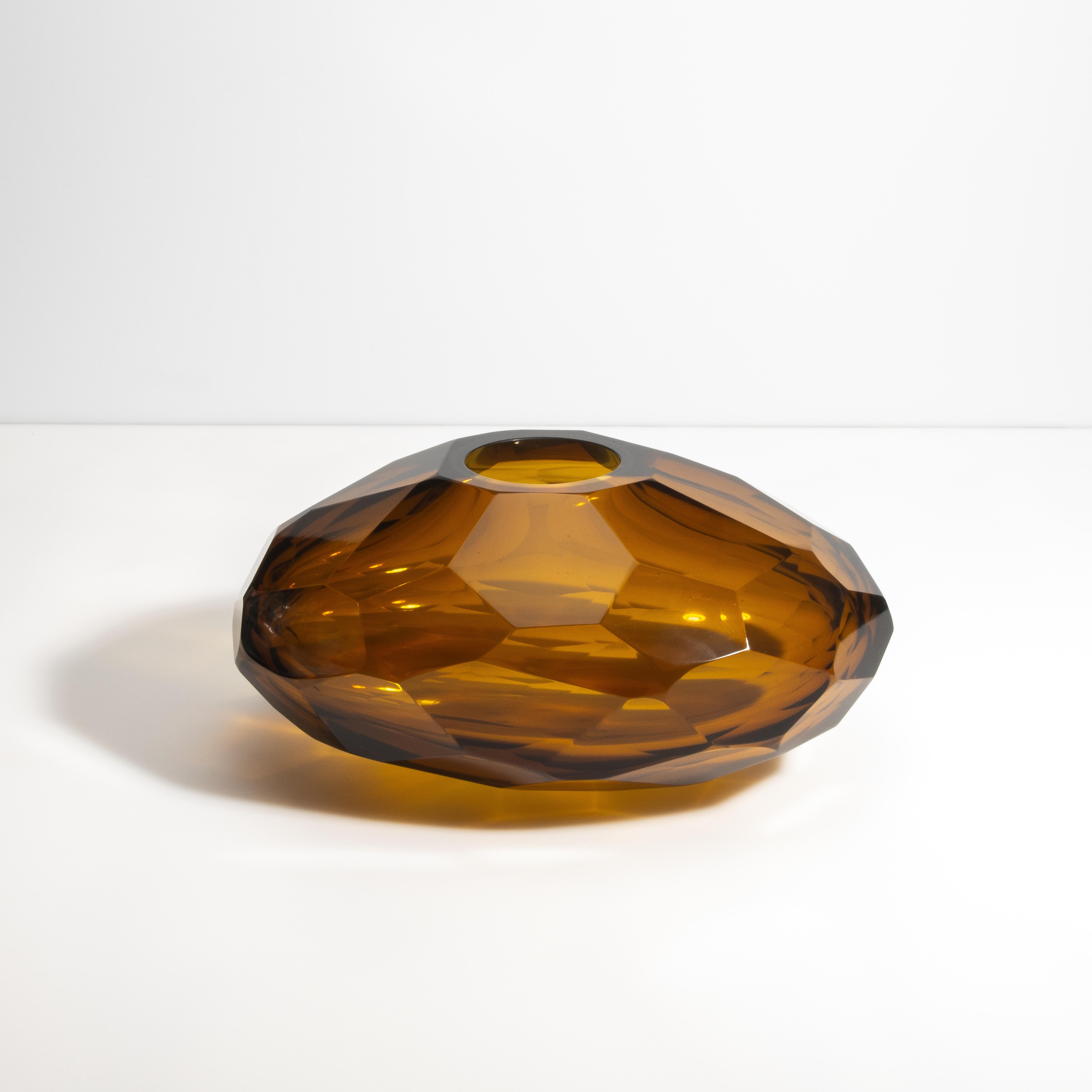 Questo squisito vaso, con la sua ricca tonalità ambrata, è una vera meraviglia di artigianato. La superficie sfaccettata crea una danza ipnotica, riflettendo un bagliore caldo e invitante che valorizza qualsiasi spazio. La sua forma geometrica