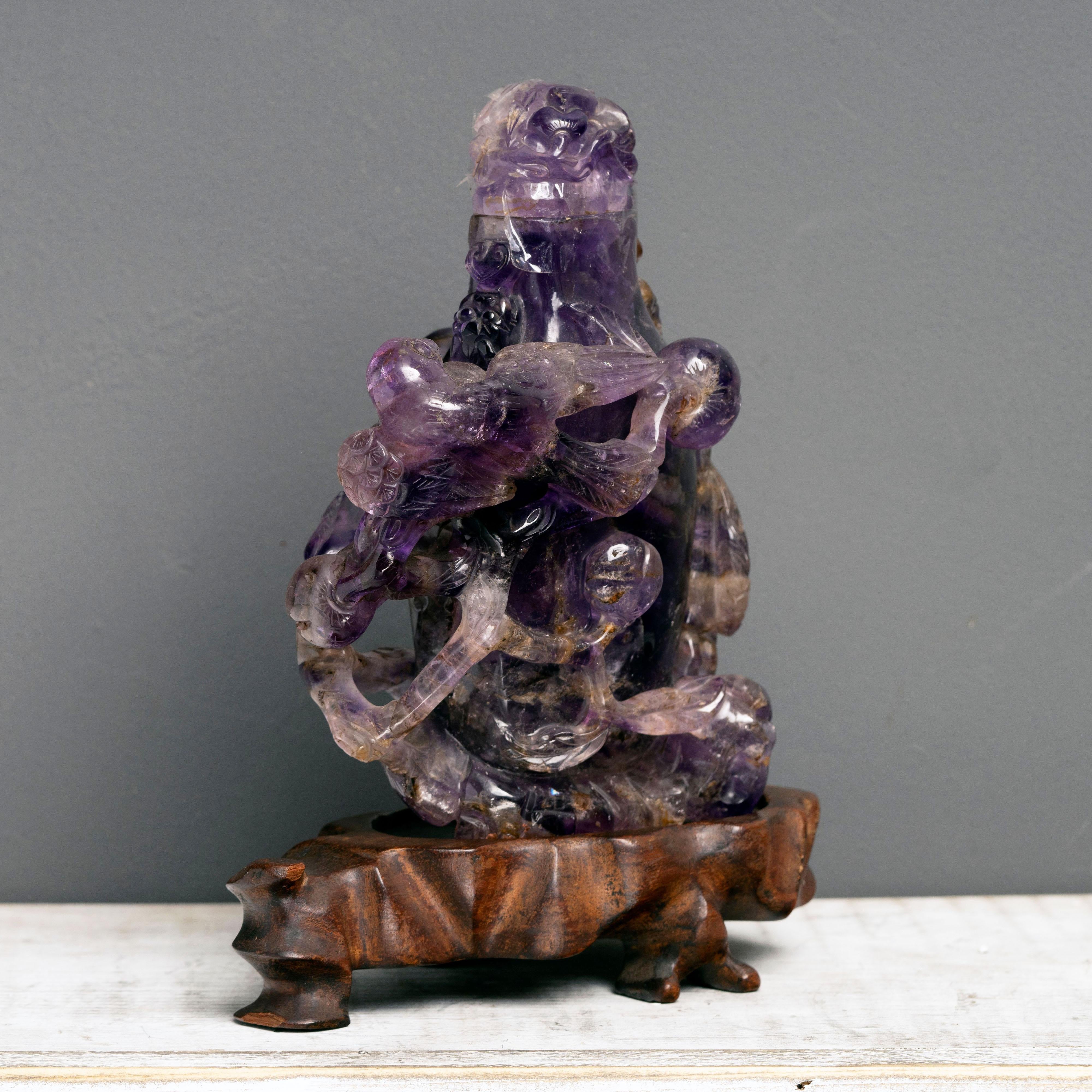 Ce superbe flacon, délicatement sculpté à la main et doté d'un bouchon amovible, est fait d'un seul morceau d'améthyste solide, gemme, d'un violet profond à clair. Il est monté sur un socle en bois sculpté sur mesure et arbore une imagerie mythique.