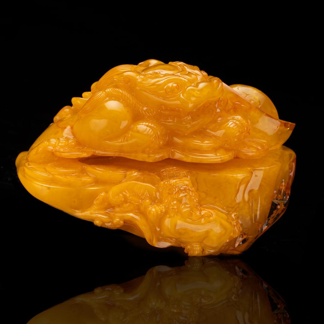 This exquisitely detailed dragon on a bed of coins and a rocky outcropping is entirely and intricately hand-carved out of one solid 245-gram piece of gorgeously-colored Baltic amber from Ukraine. This carved 34 to 55 million year specimen varies in